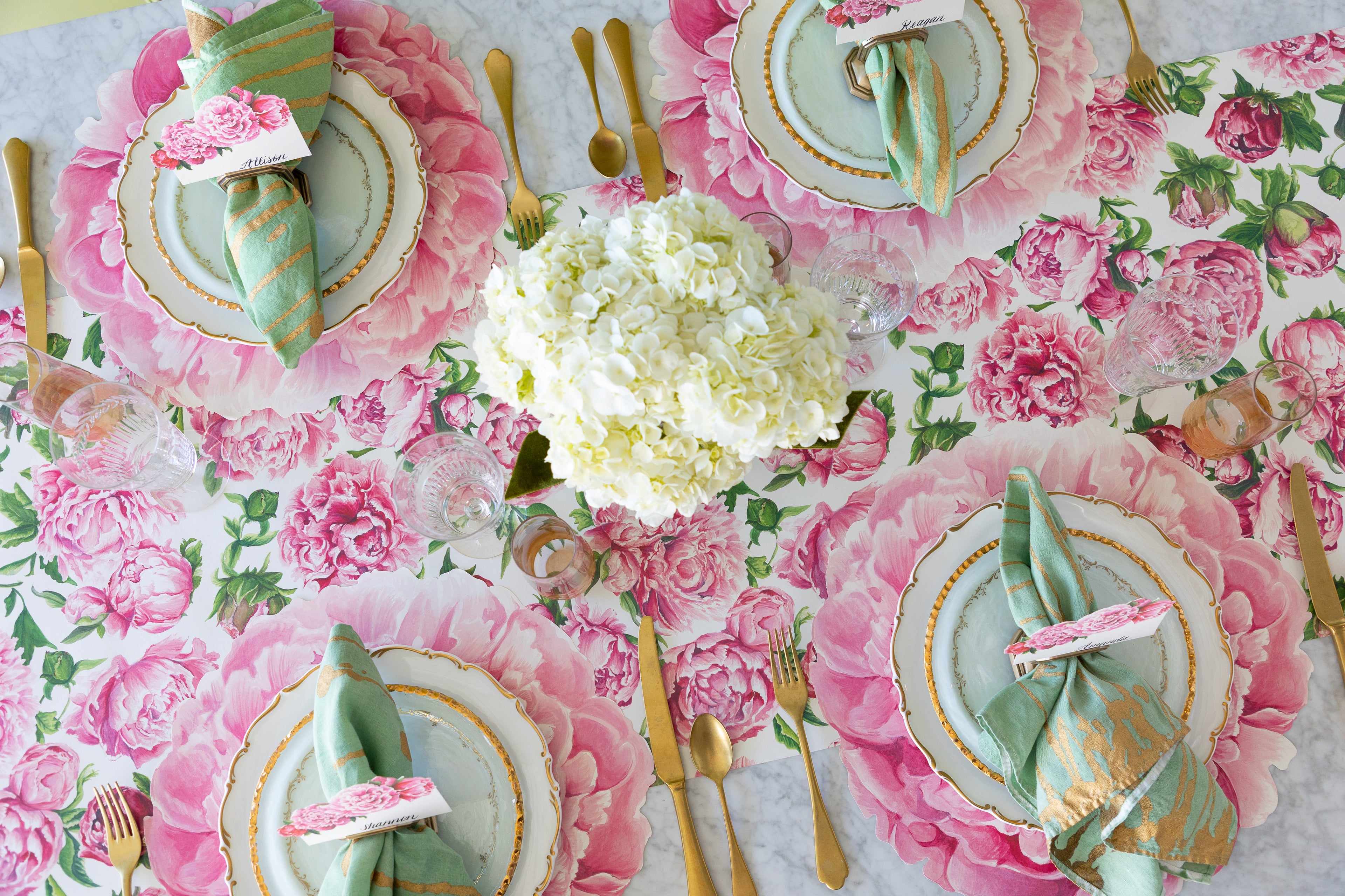 A decorative table setting with the Peonies in Bloom Runner, Die-cut Peony Placemats, decorative plates, Peonies Place Cards, marbled napkins and gold flatware.