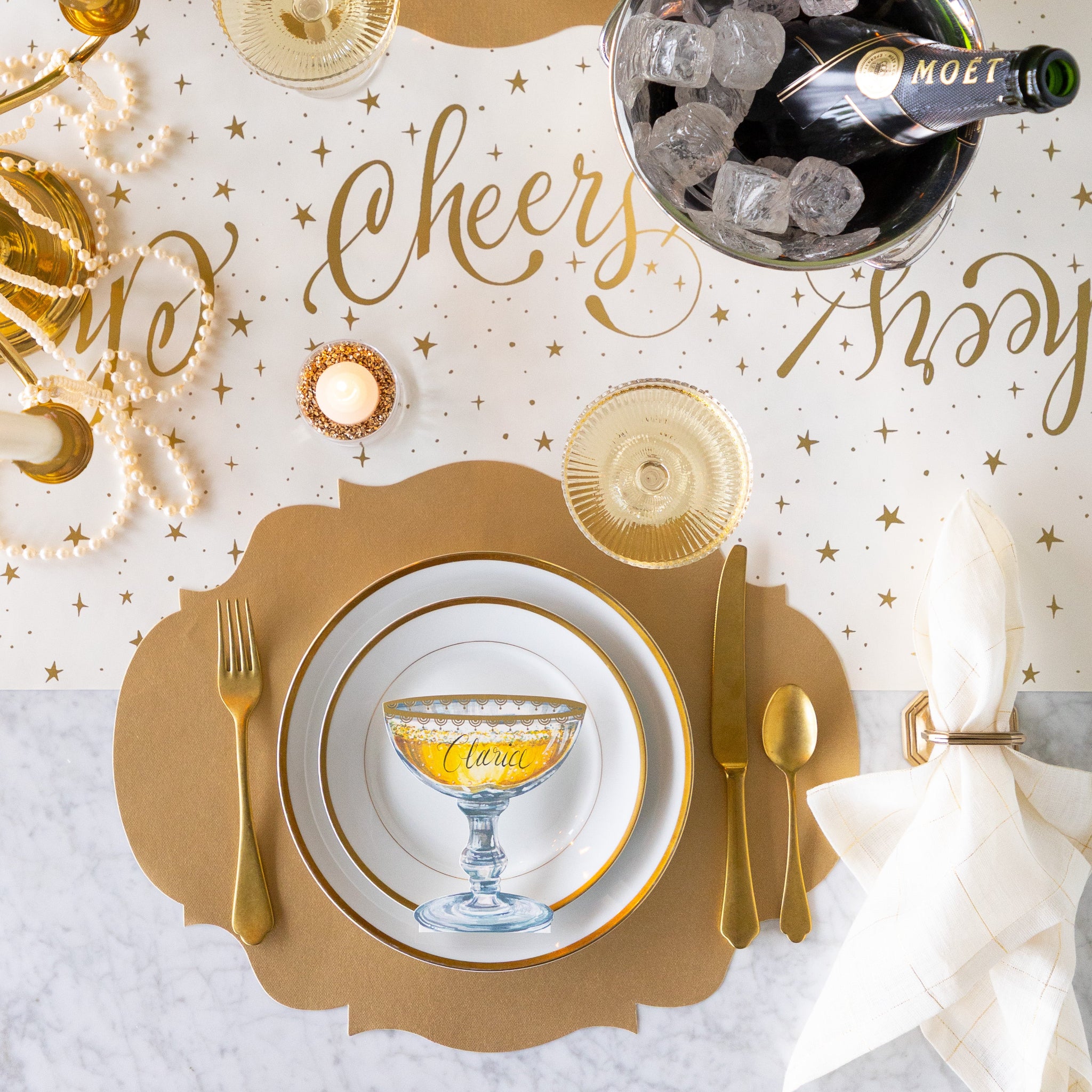 A celebratory table setting featuring an ice bucket with champagne, Die-cut Gold French Frame Placemat, gold rimmed plates, Coupe Place Card and gold flatware on the Cheers Runner.