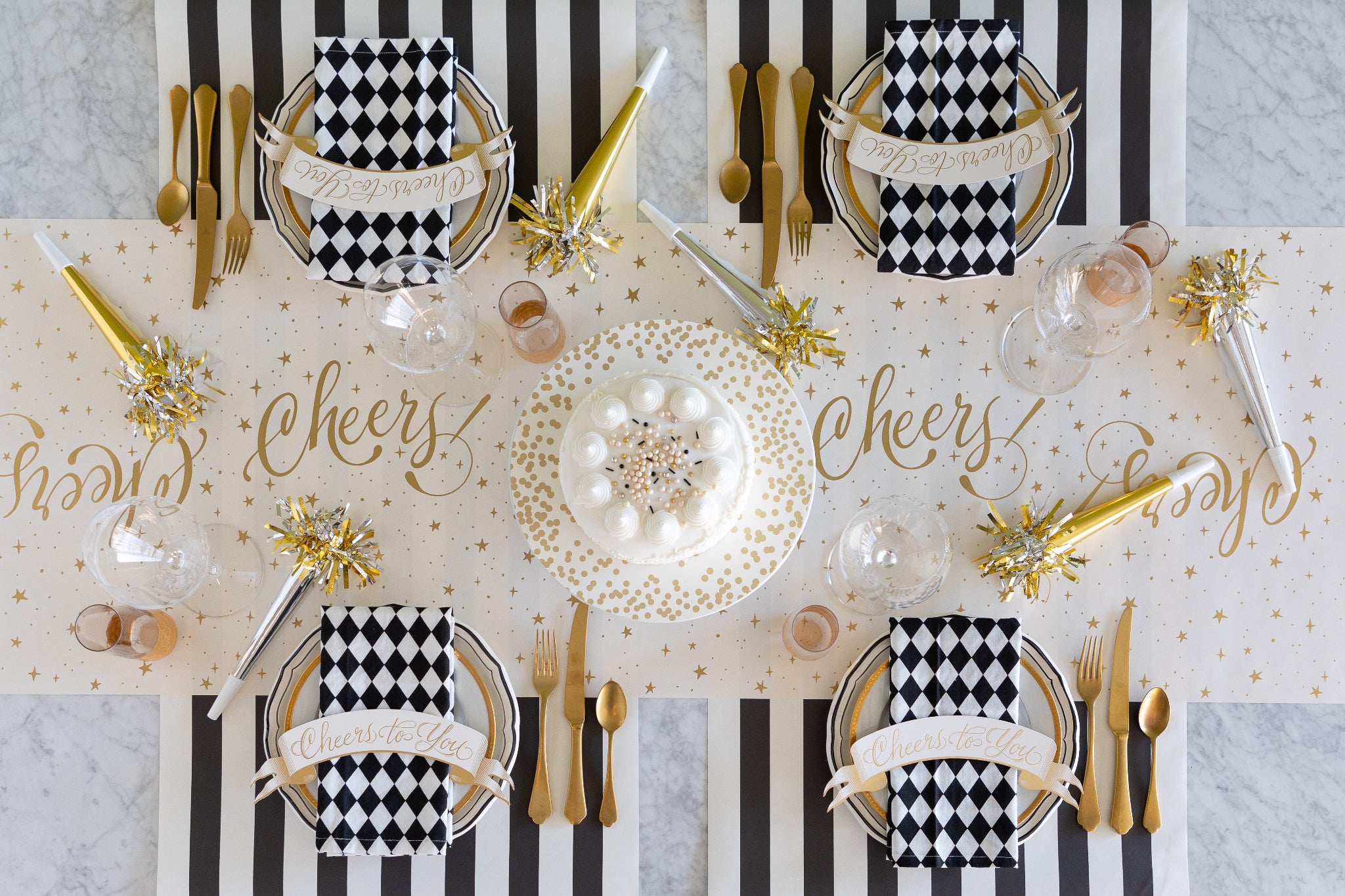 Dining table setting with black and white checkered napkins, gold cutlery, Cheers to You Banner Table Accents, party horns and cake on the Cheers and Black Classic Stripe Runners.
