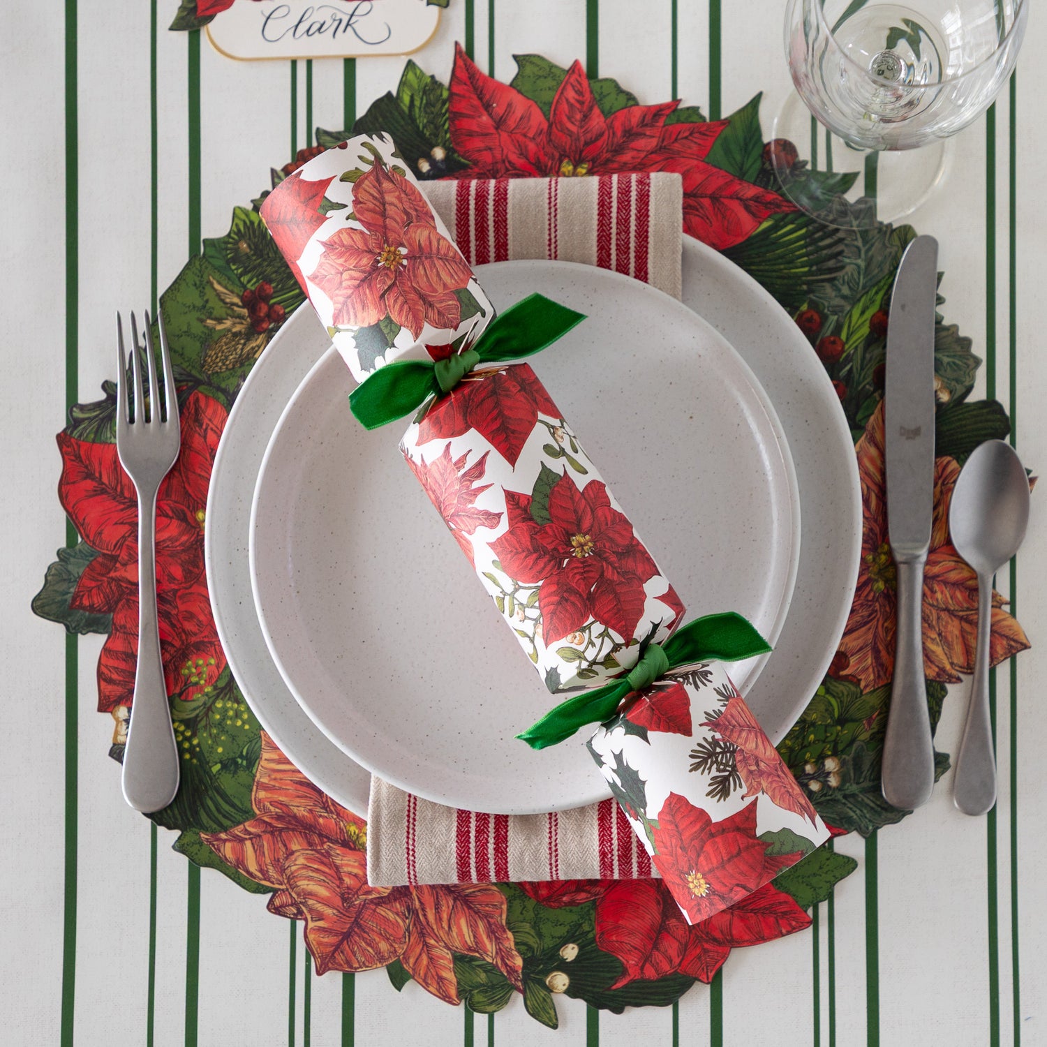 An elegant place setting featuring the Die-cut Poinsettia Wreath under white dinner plates with the Poinsettia Cracker atop, a red striped napkin, silver flatware, wine glass and Green Awning Stripe Runner underneath.
