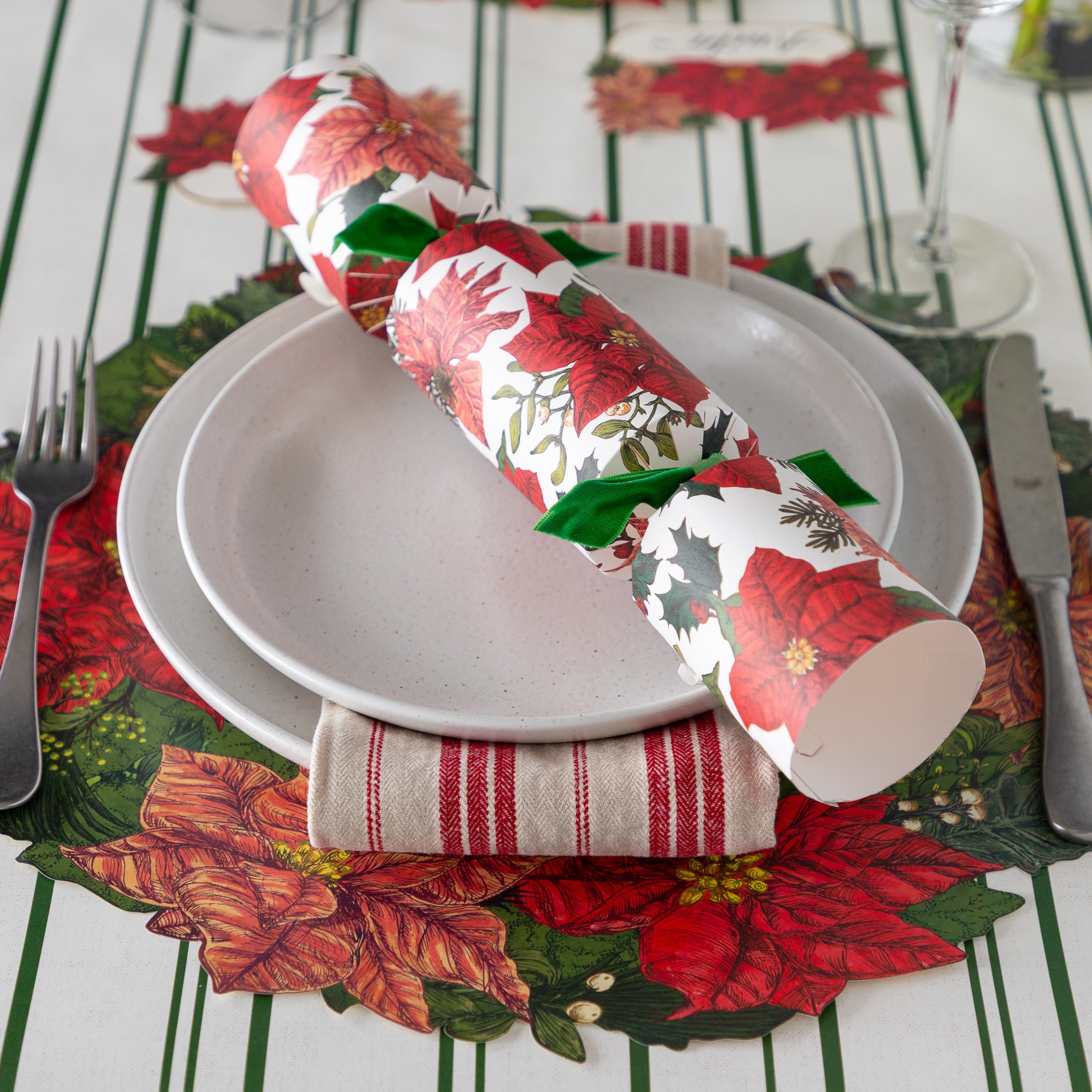 An elegant place setting featuring the Die-cut Poinsettia Wreath under white dinner plates with the Poinsettia Cracker atop, a red striped napkin, silver flatware, wine glass and Green Awning Stripe Runner underneath.
