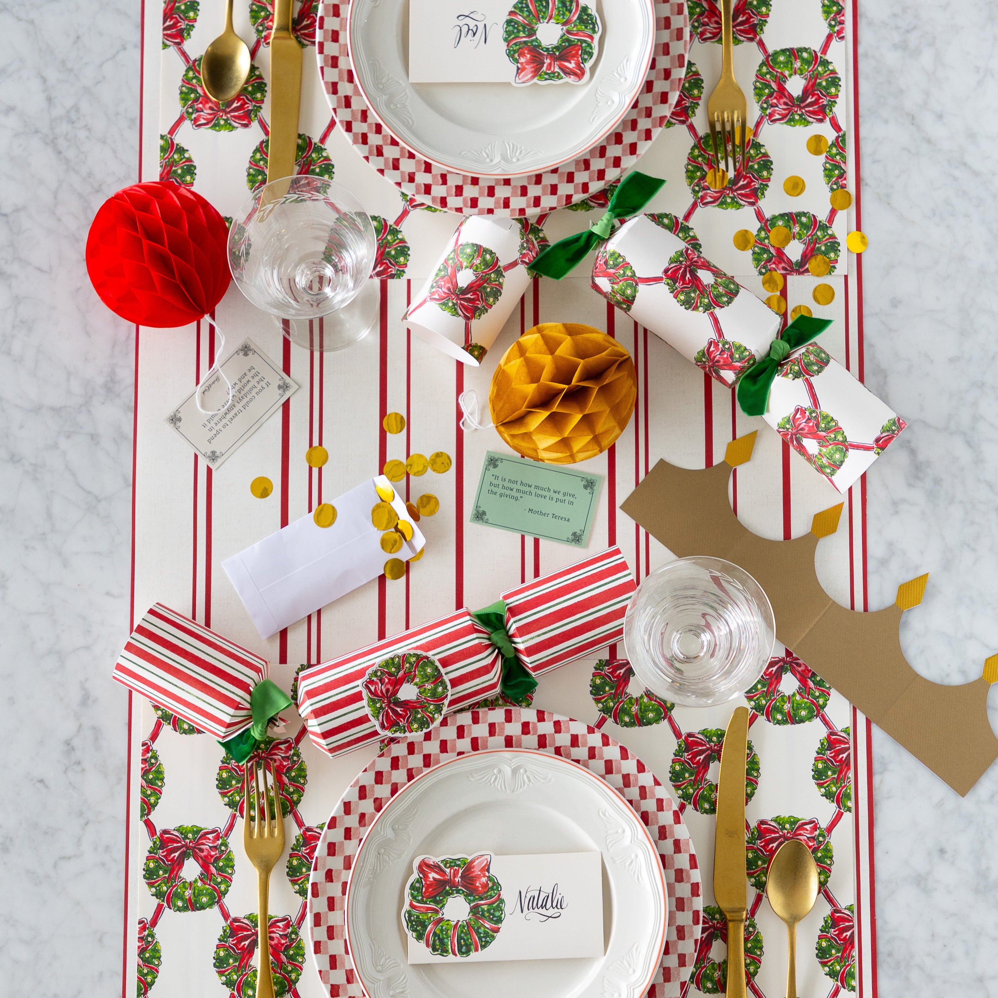 A Christmas table setting featuring the Holiday Wreath Placemats, Holiday Wreath Place Cards atop dinner plates, Holiday Wreath Crackers partially opened with their contents on the table, and Red Awning Stripe Runner underneath.