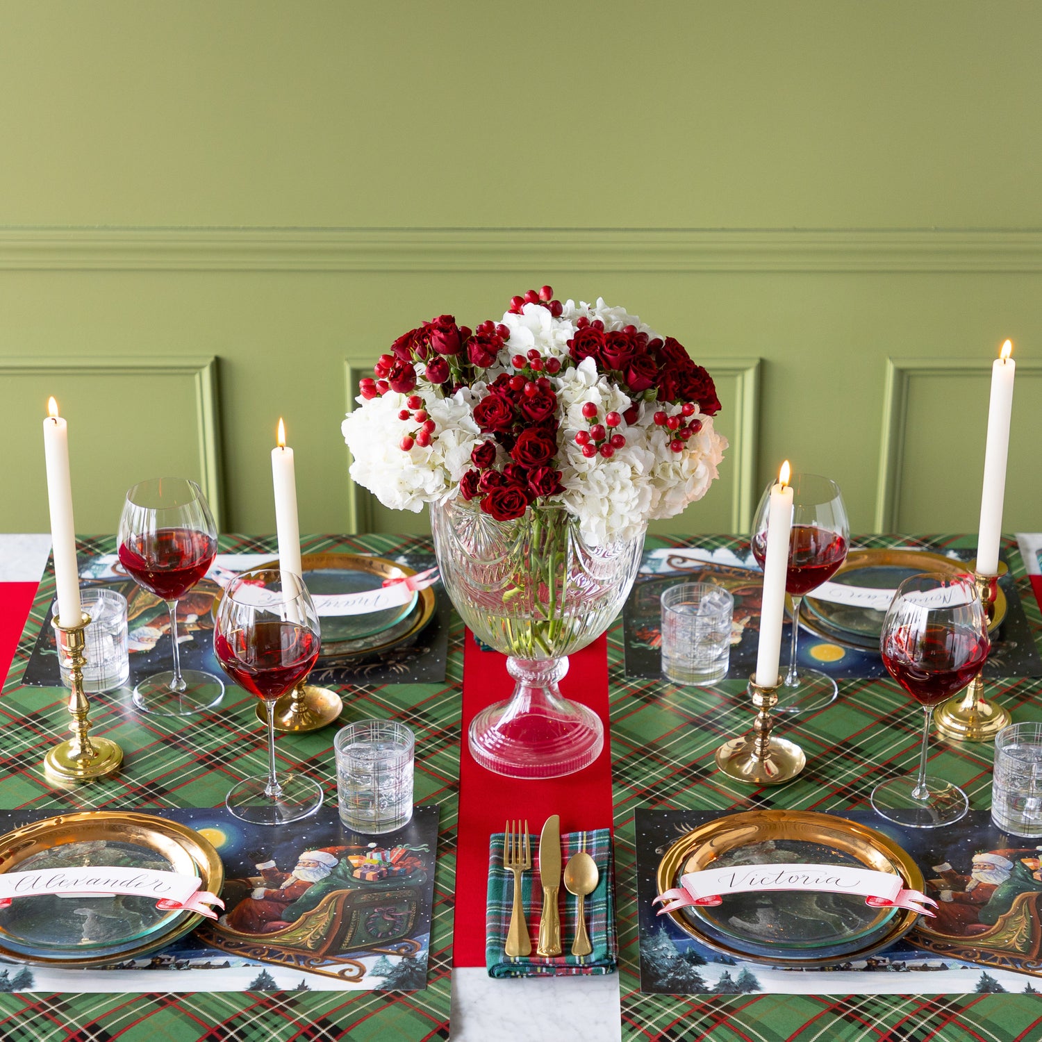A Christmas table setting with a red and white floral centerpiece, featuring the Sleigh Ride Placemats, gold rimmed dinnerware, lit candles, glasses of wine and water, Green Plaid Runner rolled vertically underneath and Red Flocked Runner rolled horizontally underneath.