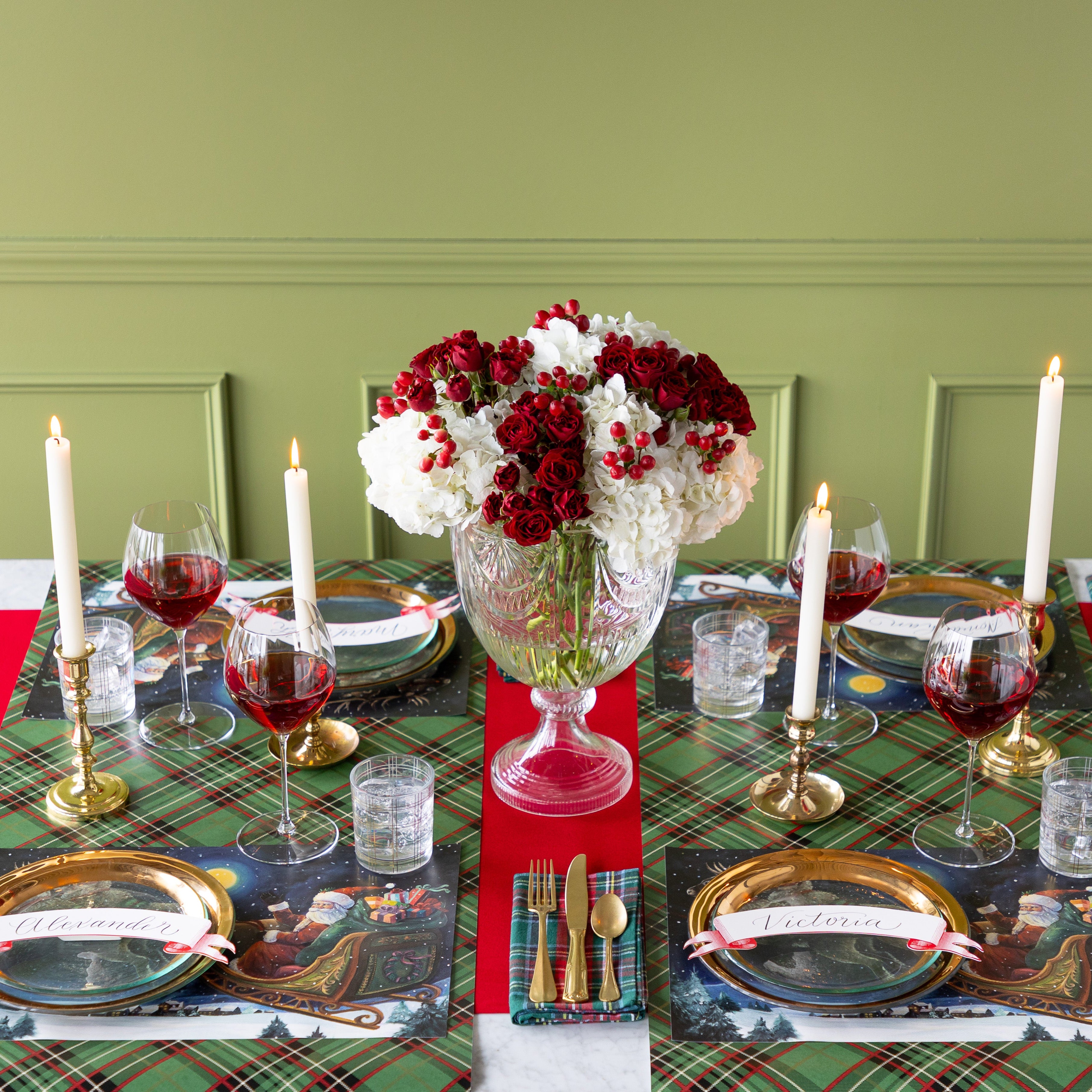 A Christmas table setting with a red and white floral centerpiece, featuring the Sleigh Ride Placemats, gold rimmed dinnerware, lit candles, glasses of wine and water, Green Plaid Runner rolled vertically underneath and Red Flocked Runner rolled horizontally underneath.