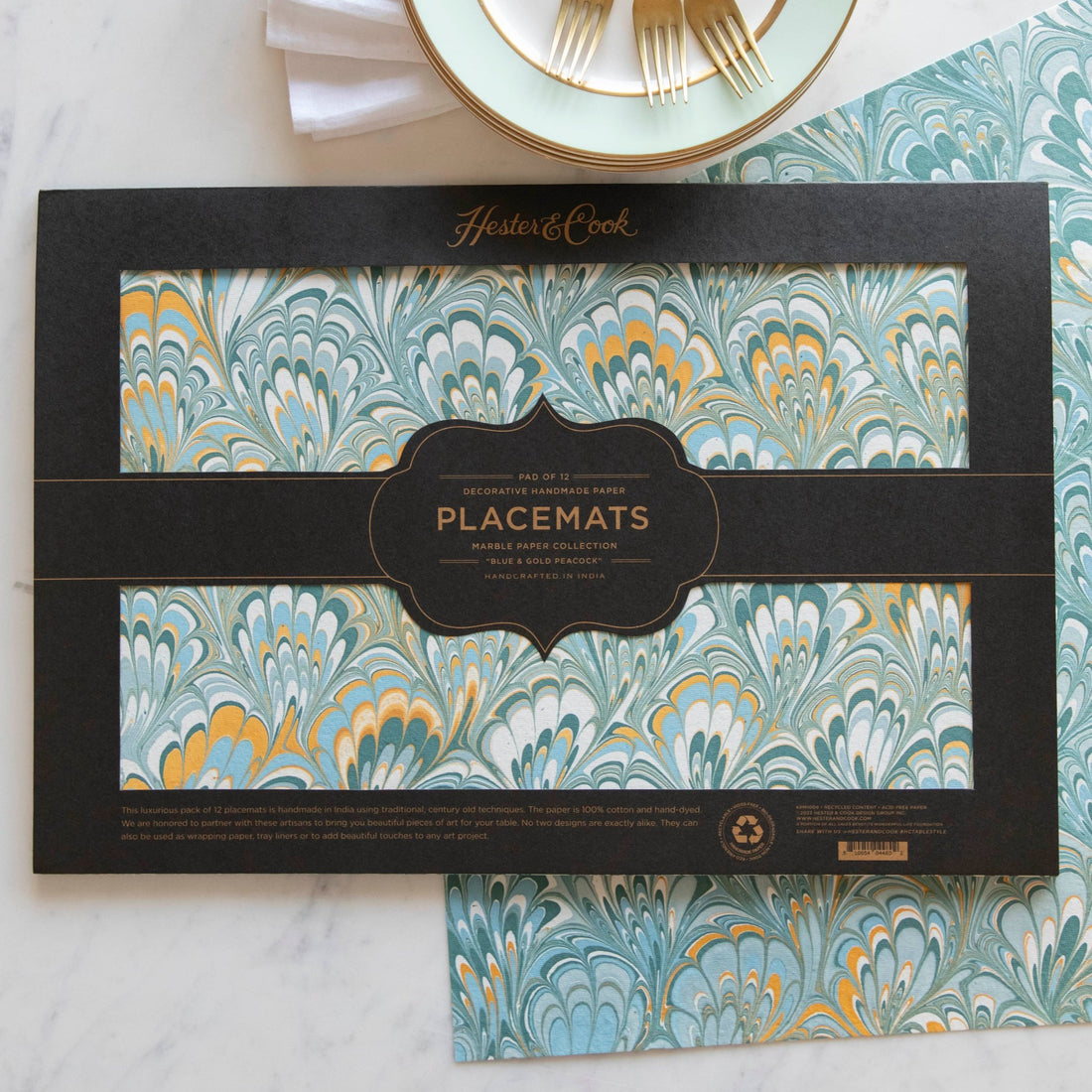 The Blue &amp; Gold Peacock Marbled Placemat in the black, open-face packaging, with two placemats on the table beneath.