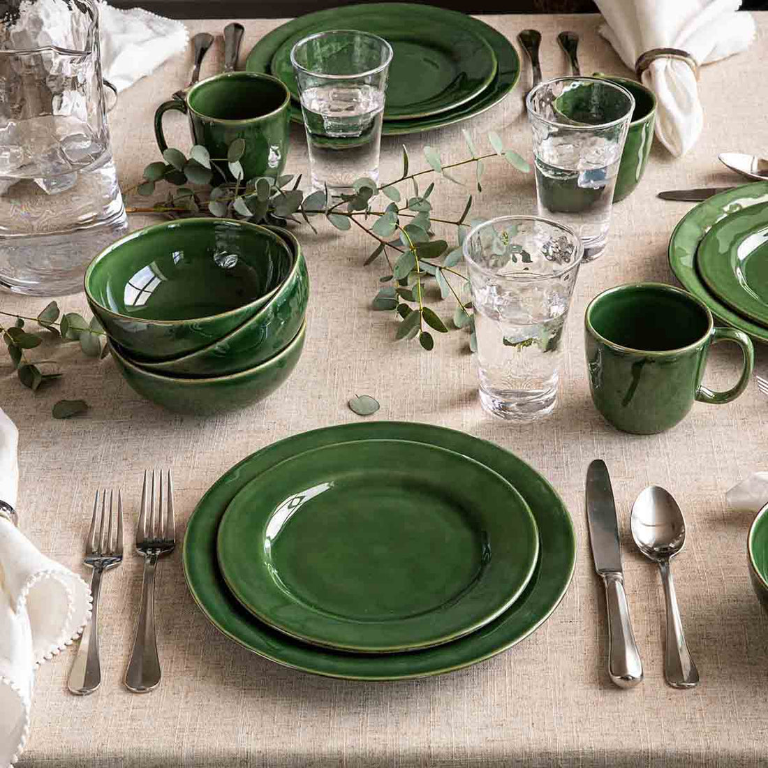 A table setting with Juliska Puro Basil Dinnerware dishes, silverware, glasses, and a natural greenery decoration.