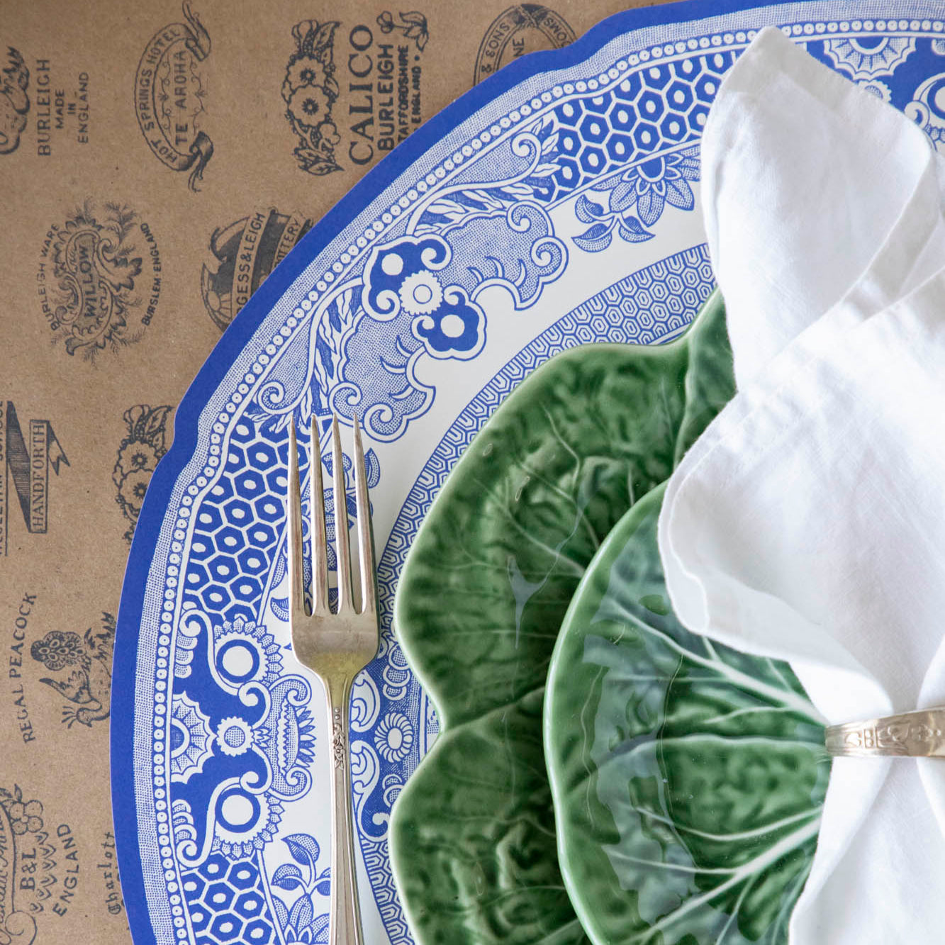 Die-cut Blue Willow Plate Placemat with an elegant place setting on top and Kraft Burleigh Heritage Runner underneath.