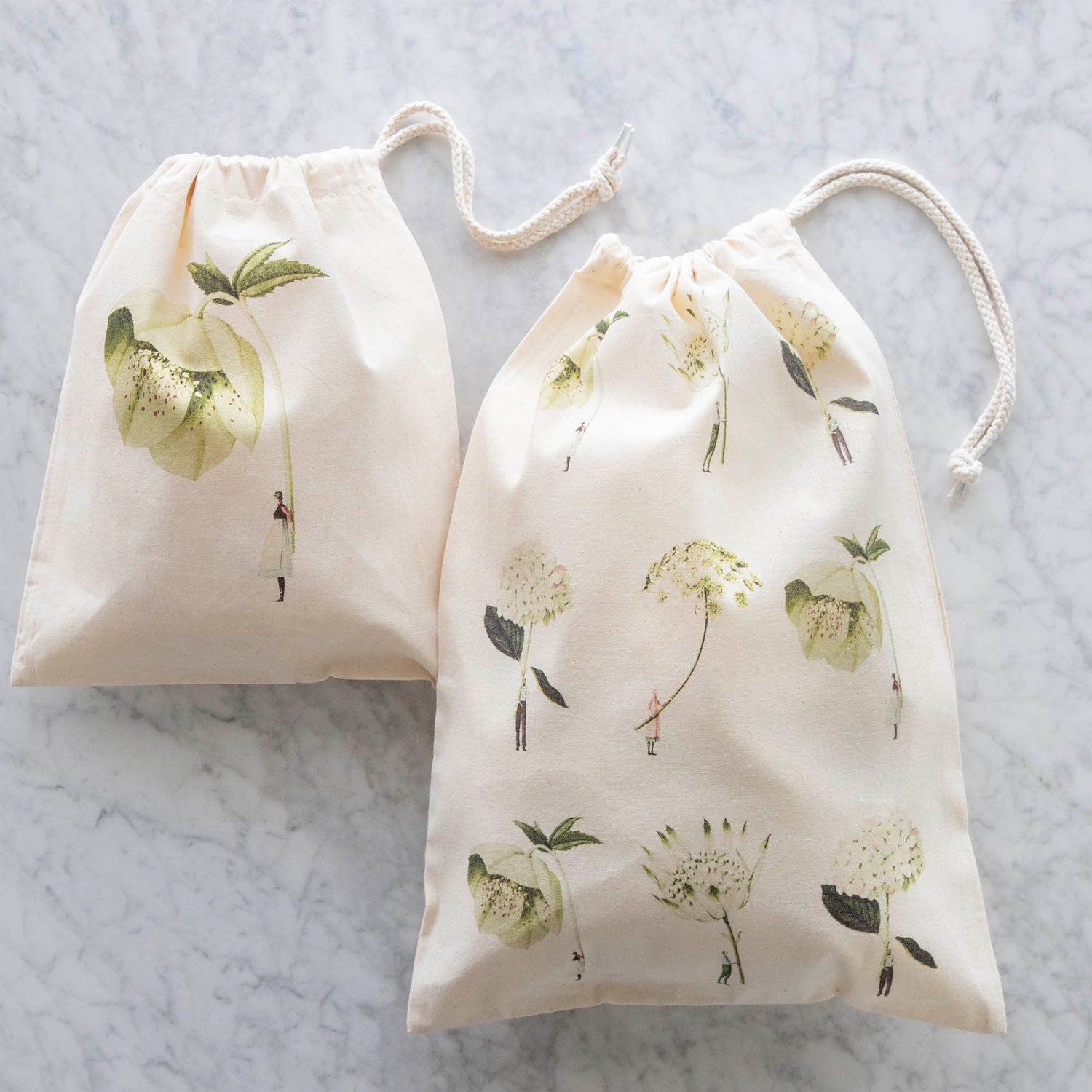 Two Laura Stoddart Drawstring Bags on a white table - the Small Hellebore Bag and the Medium Green Flowers Bag, both cinched closed. 