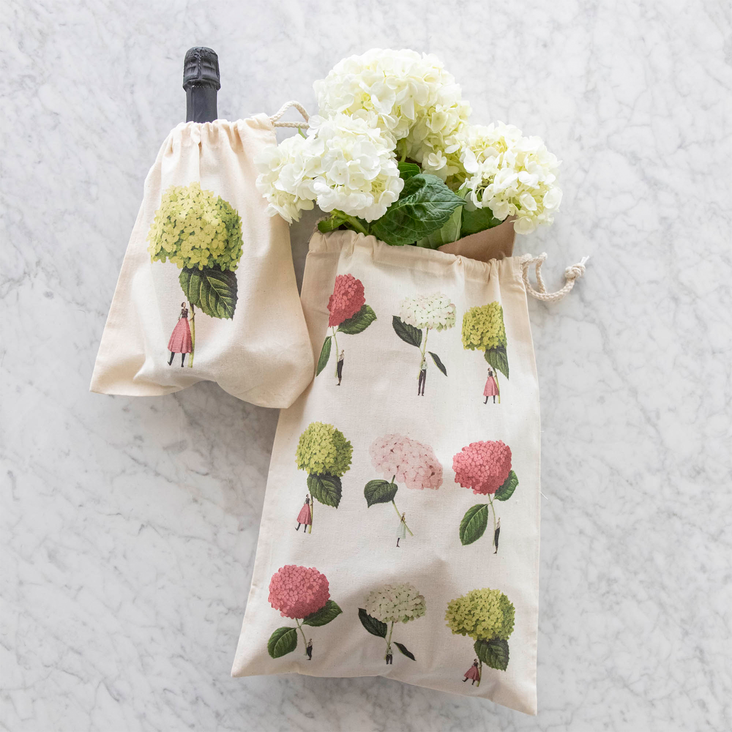 Two of the Laura Stoddart Drawstring Bags resting on a white table - the Small Anabelle Bag containing a bottle of wine, and the Medium Hydrangeas Bag containing white blooms. 