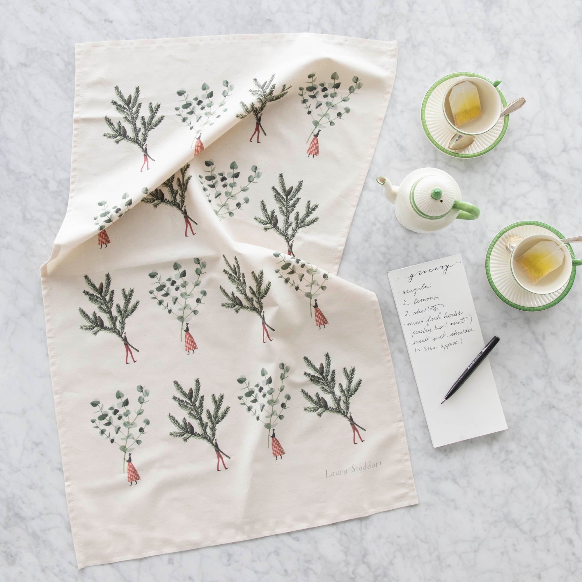 Laura Stoddart Spruce &amp; Eucalyptus Tea Towel next to a notepad with a grocery list written on it, and tea cups and a creamer pot.