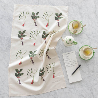 The Laura Stoddart Holly &amp; Mistletoe Multi Tea Towel next to a grocery list, tea cups and a creamer pot.