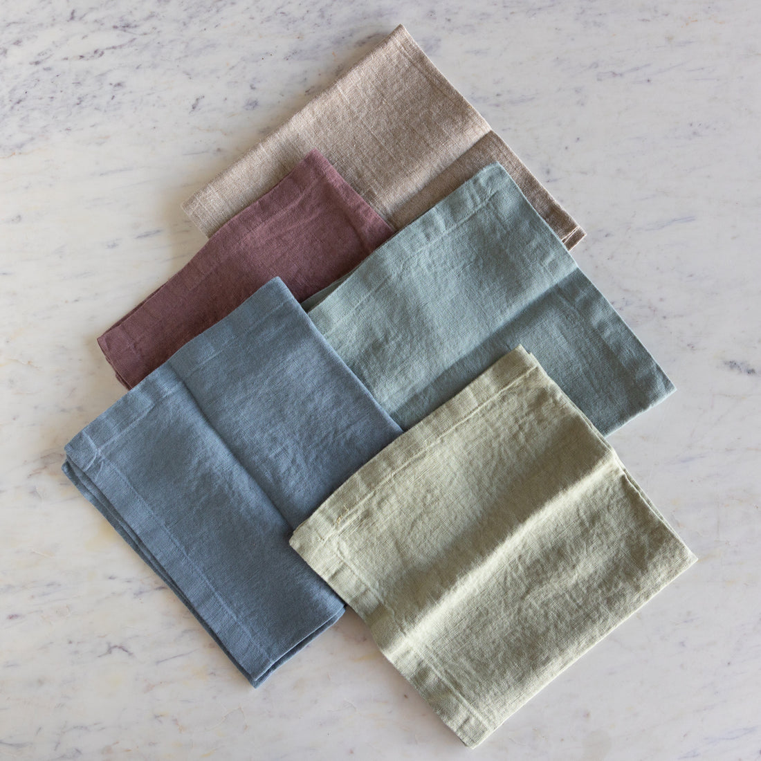 Set of five folded napkins in various earthy tones on a light marble surface.