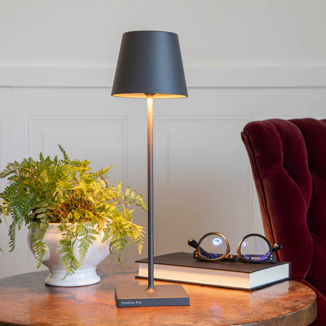A touch-dimming control Zafferano Dark Grey Cordless Lamp on a table next to a book and a plant.