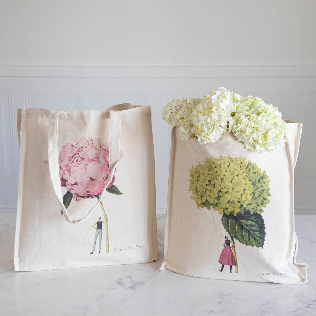 Two Hester &amp; Cook heavyweight canvas tote bags with floral prints leaning against wooden crates beside potted topiary plants.