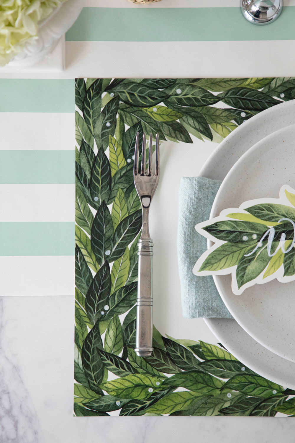 A beautifully arranged place setting with a fork and a plate, perfect for holiday parties or weddings. The table is elegantly decorated with the Hester &amp; Cook Laurel Placemat, adding a touch of sophistication to.