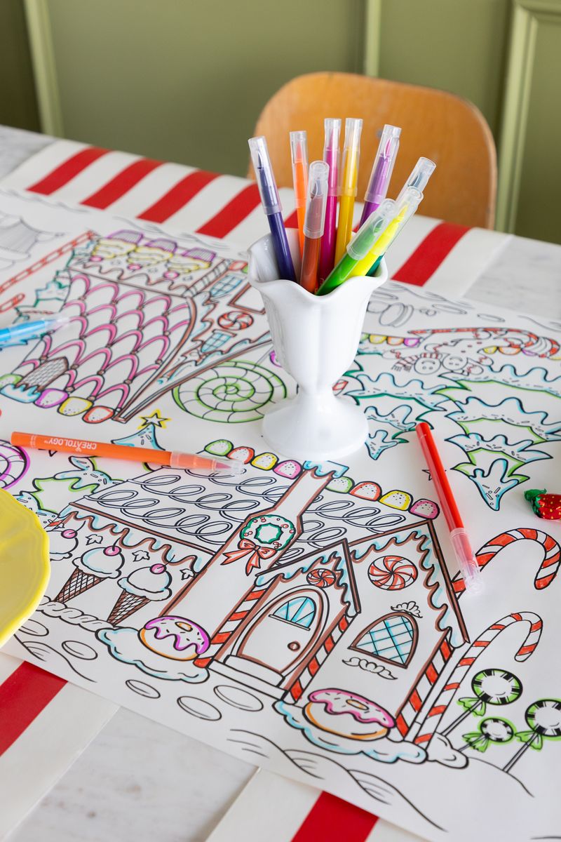 A paper coloring runner with a partially colored in gingerbread house on a table with colorful markers. 