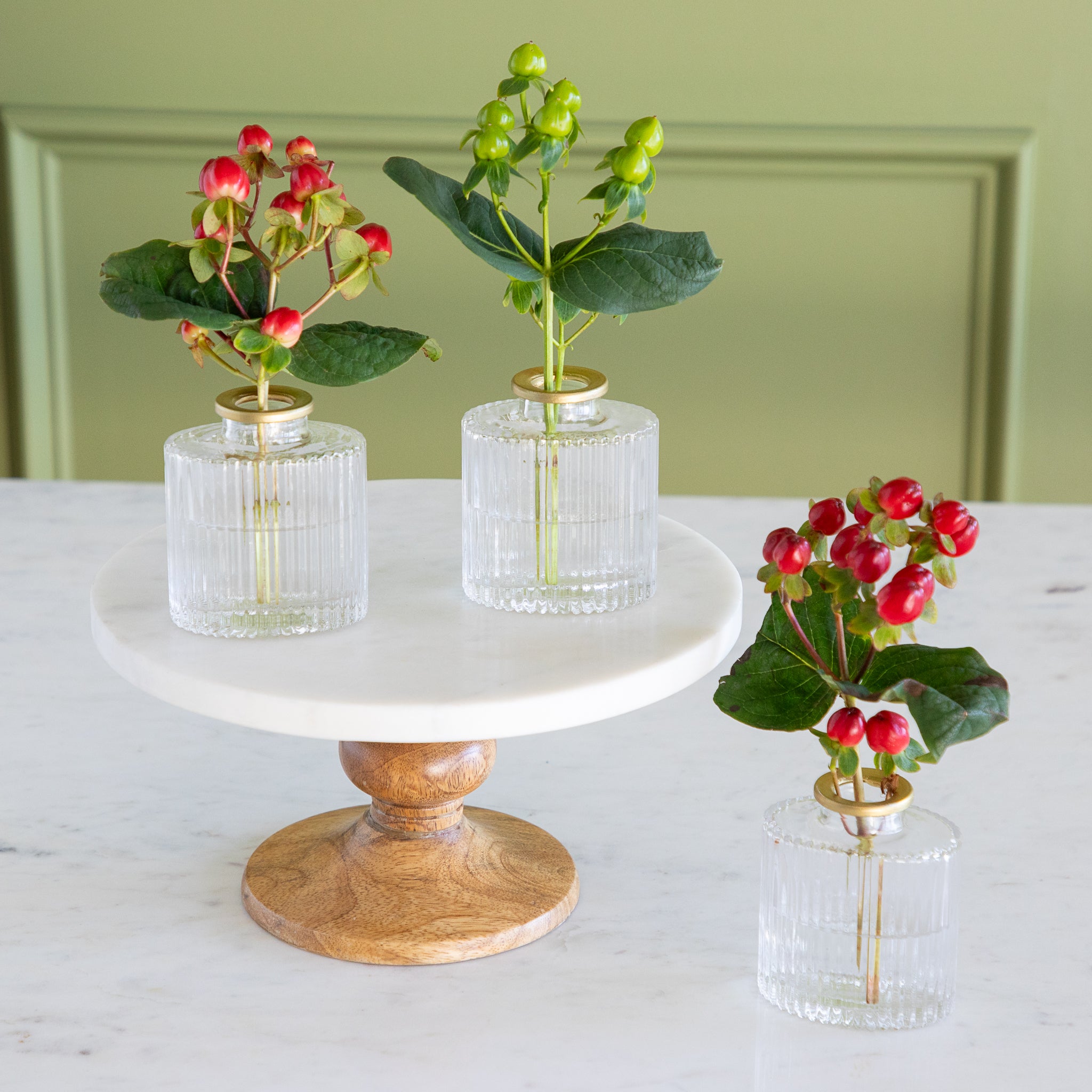 Three small plants in glass vases on a marble and wood stand against a light green wall.