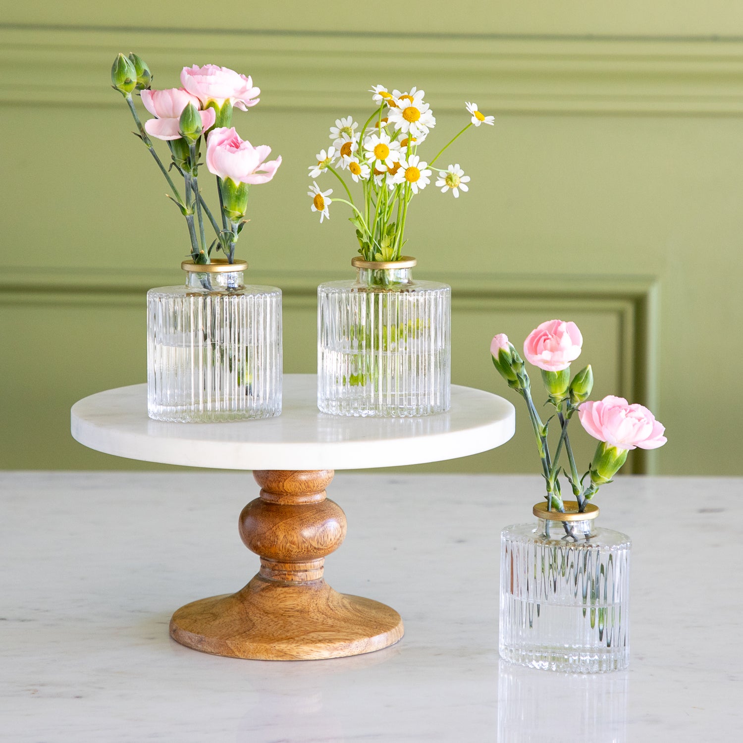 Three small glass vases with flowers on a marble and wood stand against a green wall.