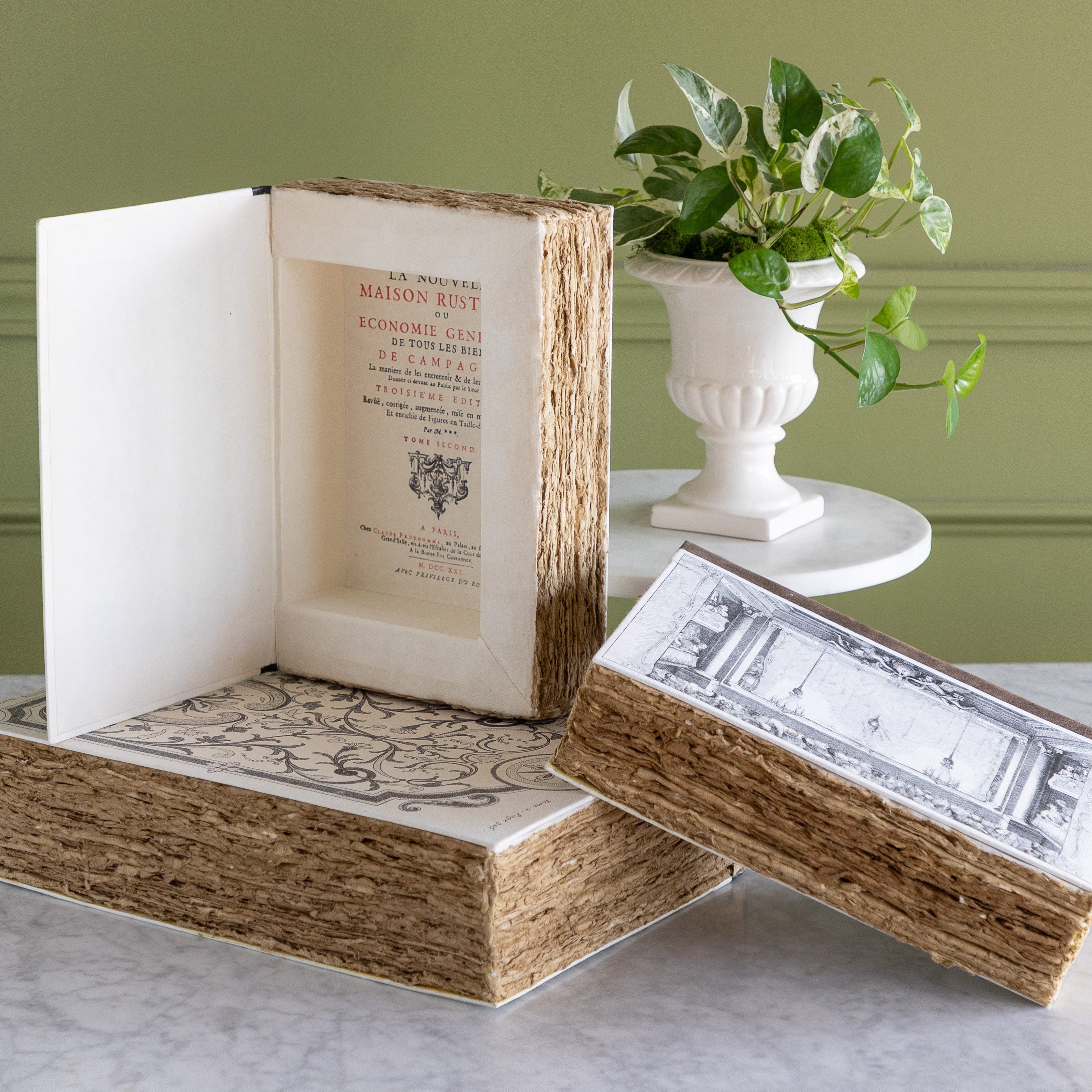Decorative storage boxes resembling vintage books displayed on a marble table with a plant behind.