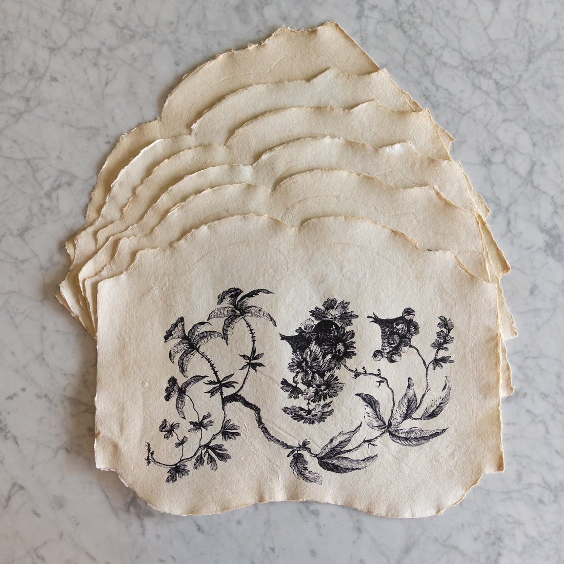 Stack of decorative paper placemats with floral and bird design on a marble surface.