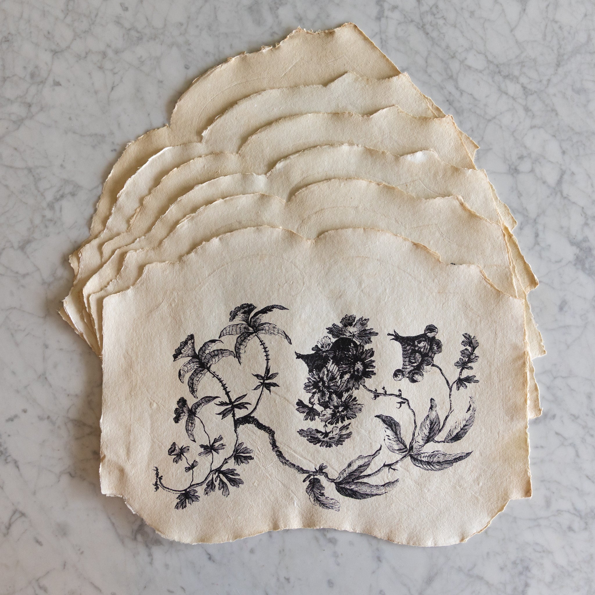 Stack of decorative paper placemats with floral and bird design on a marble surface.