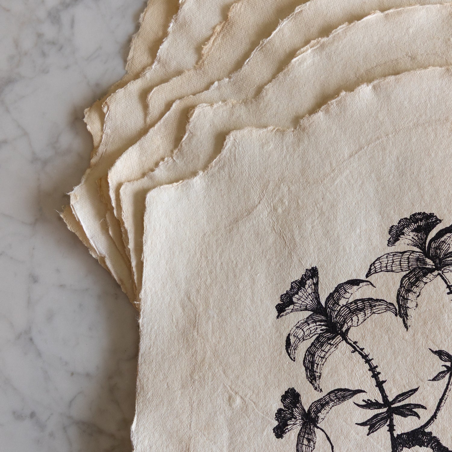 Stack of textured paper placemats with black floral design on a marble surface.