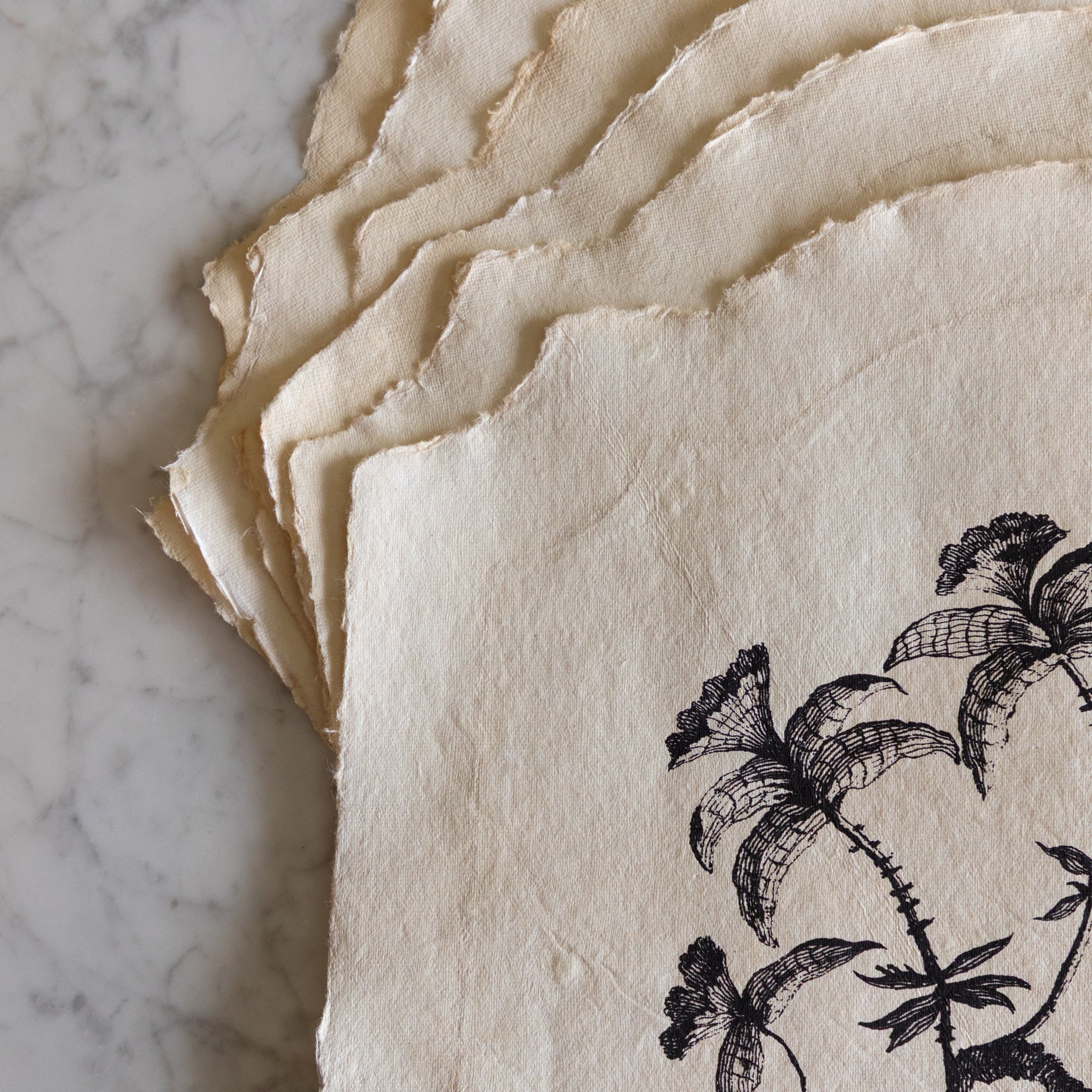 Stack of textured paper placemats with black floral design on a marble surface.
