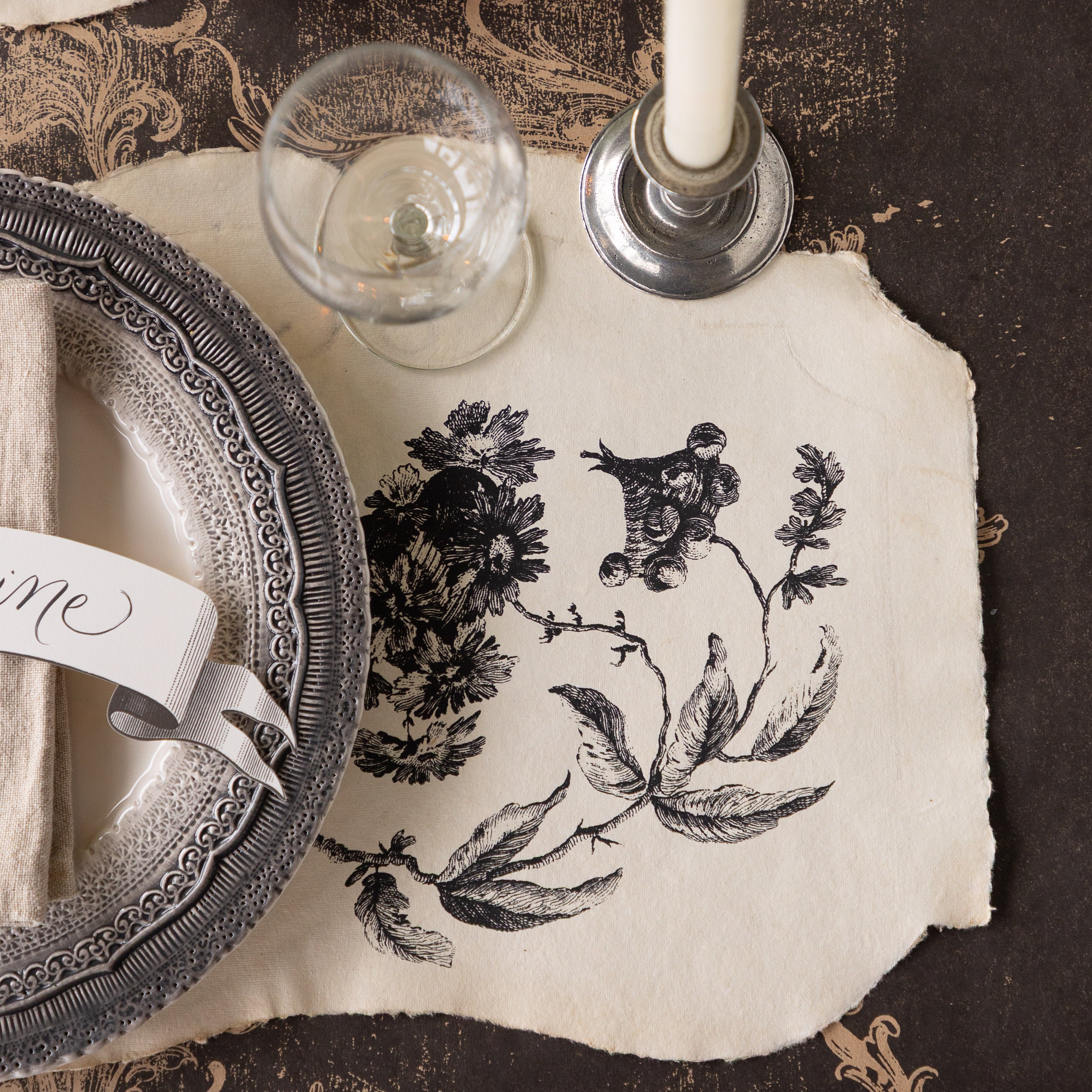 Elegant place setting with decorative plates, Pillement Paper Placemats, Classic Banner Table Accent, glasses, and candles on the Black Archival Runner.