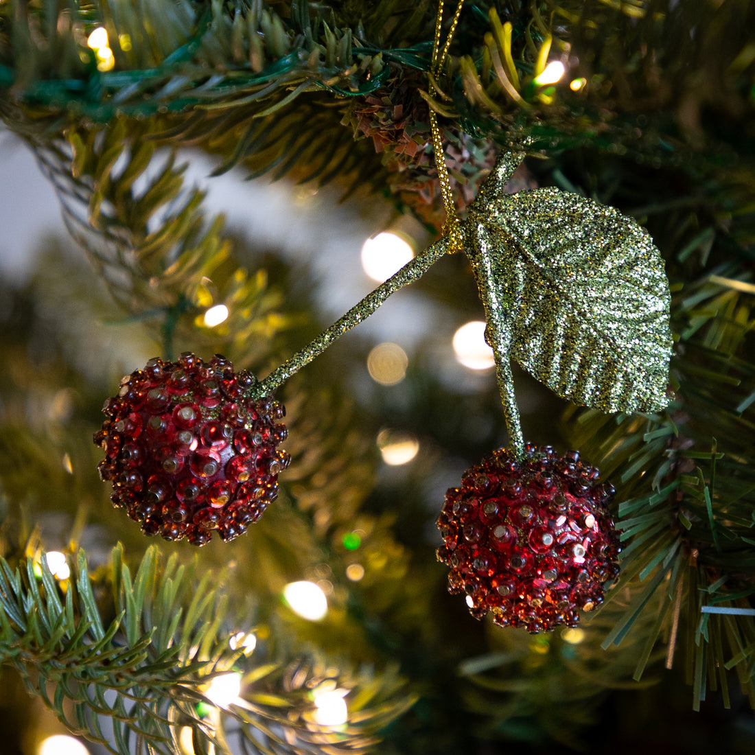 A close-up of a Christmas tree branch with a red beaded cherry ornament hanging from gold glitter stems and leaves, with warm white string lights blurred in the background.