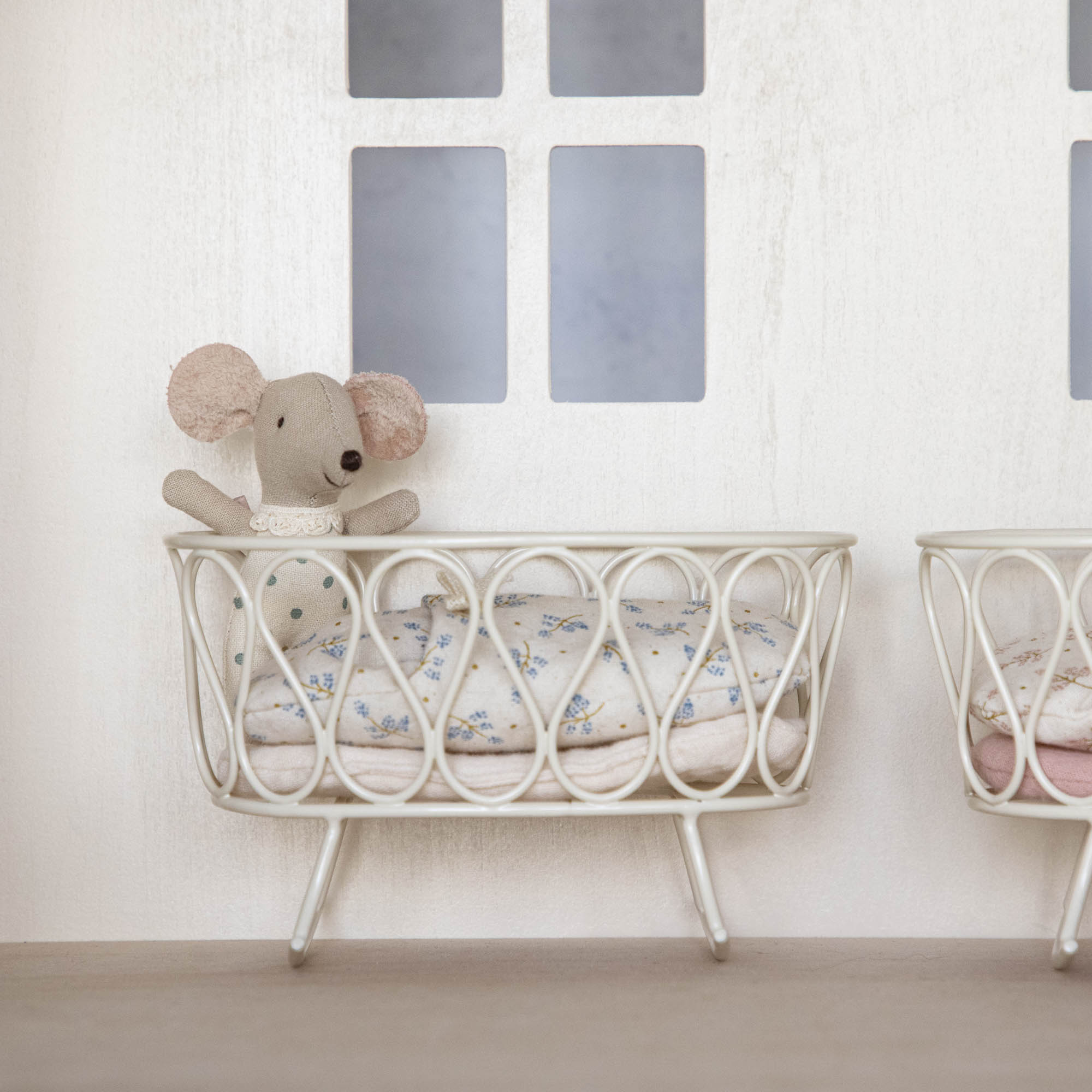 A Maileg stuffed rabbit toy resting on a white Maileg metal cradle against a wall with simple decor.