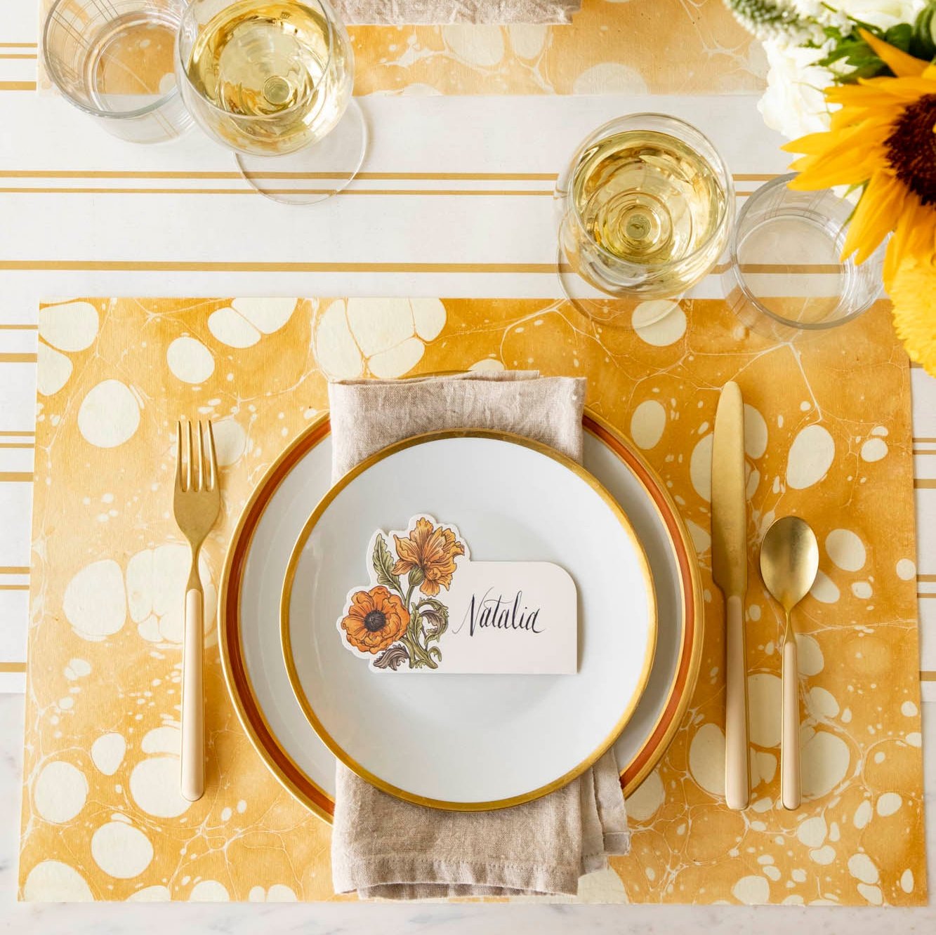 An elegant fall themed table setting with the Gold Stone Marbled Placemat, orange and gold rimmed dinner plates with the Autumn Arrangement place card with the name &quot;Natalia&quot; written on it, on top. 