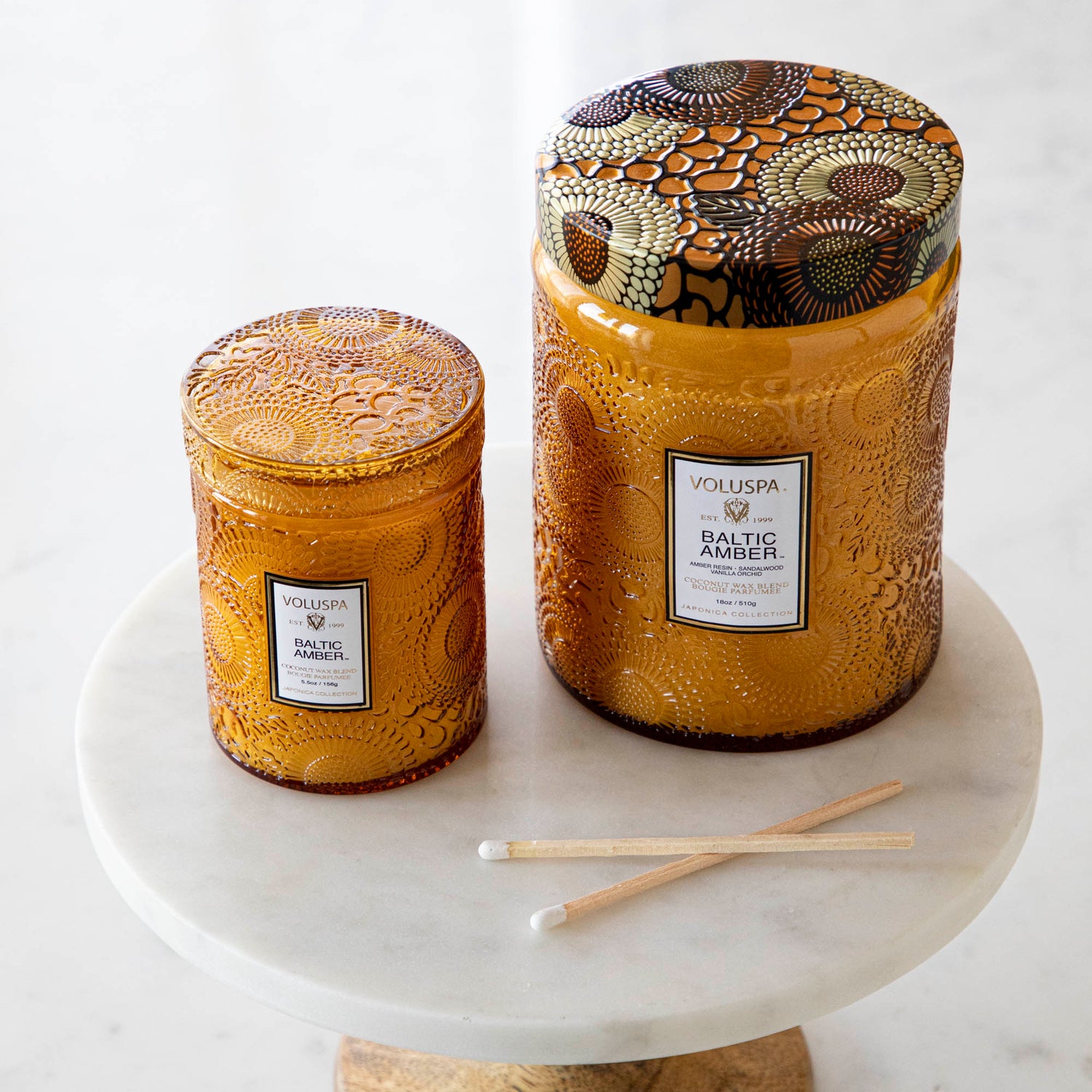 Two Voluspa Baltic Amber candles with matches on a marble surface.