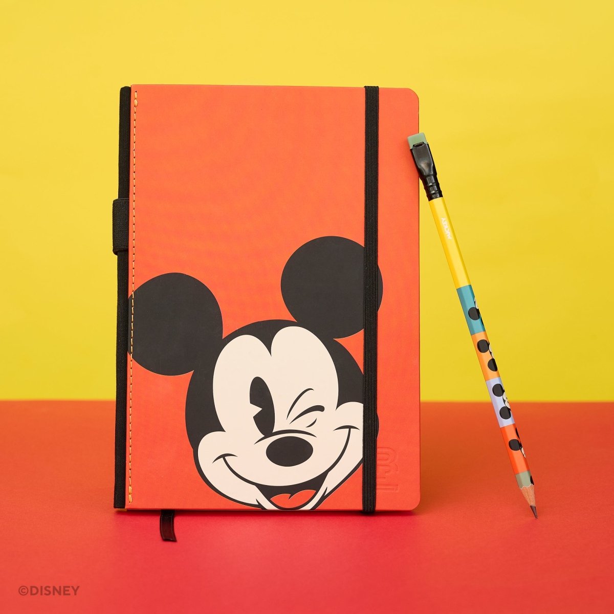 An orange Disney Mickey Mouse notebook with a black elastic closure, standing next to a yellow and blue Mickey-themed pencil on a red and yellow background.