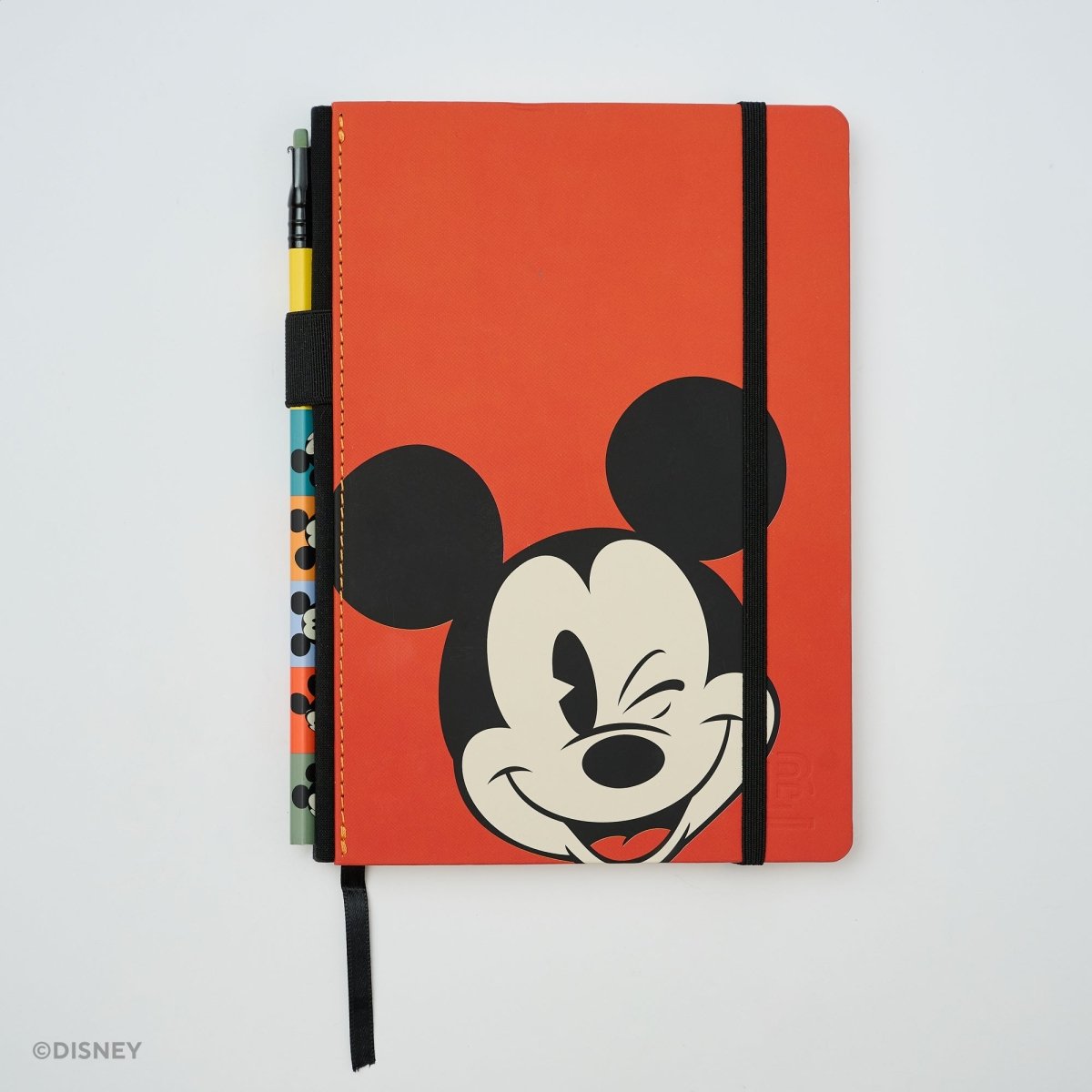 A red Mickey Mouse notebook with a black elastic closure band, featuring a winking Mickey face on the cover and a pen loop with colorful pens visible on the side, photographed on a light gray background.