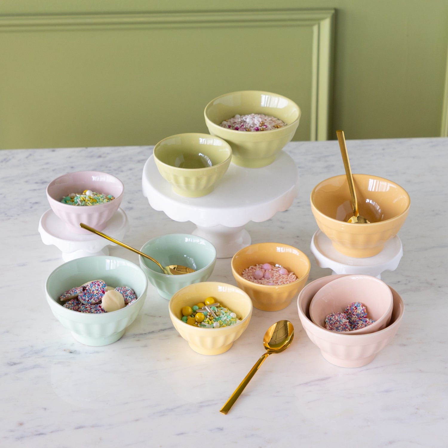 Set of colorful ceramic bowls in two sizes filled with various sprinkles and gold spoons on a marble stable.