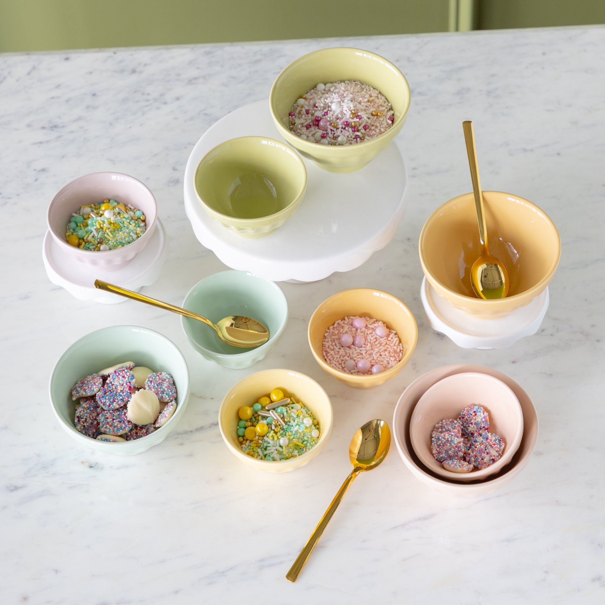 Set of colorful ceramic bowls in two sizes filled with various sprinkles and gold spoons on a marble stable.