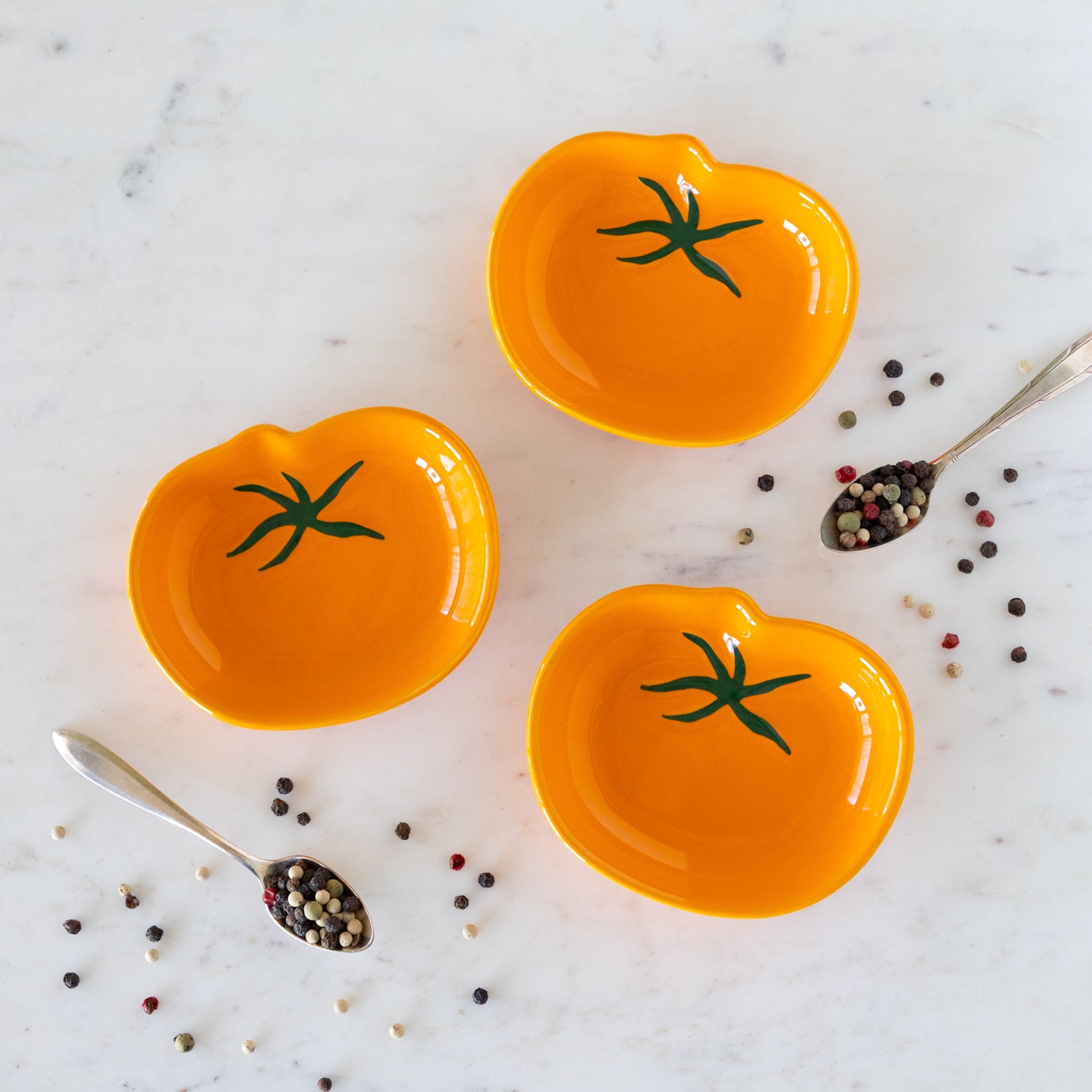 Three orange ceramic dishes shaped like tomatoes on a marble surface with peppercorns.