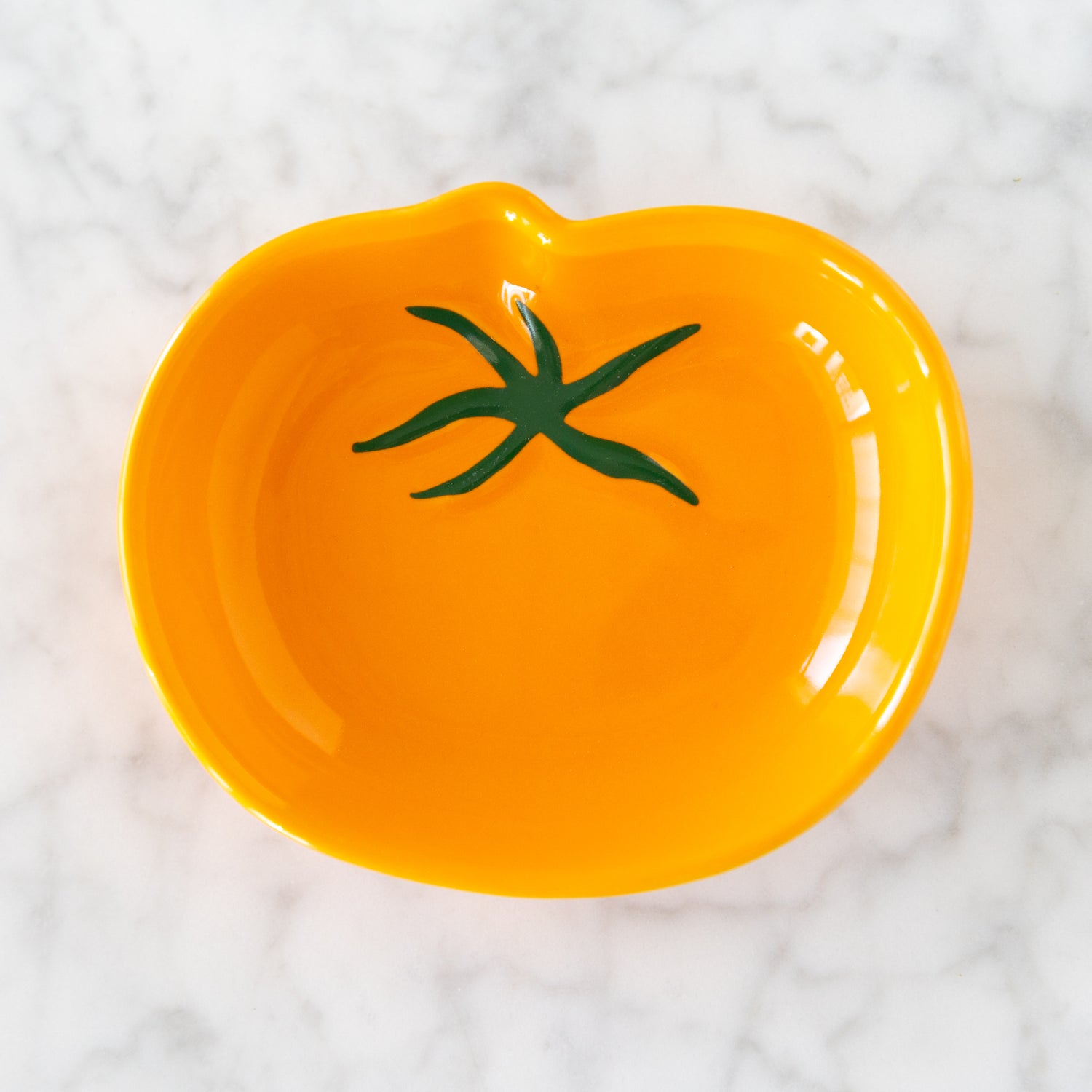 Yellow ceramic plate shaped like a tomato with green stem on a white marble background.