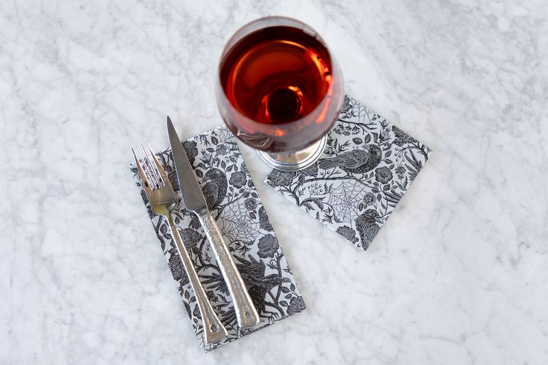 Wine glass with cutlery on a pair of silver and black Halloween themed napkins on a white marble surface.