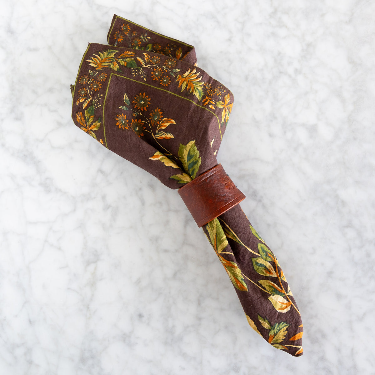 A brown patterned napkin with a leaf and floral print, tied with a matching brown leather napkin ring, displayed on a white marble surface.