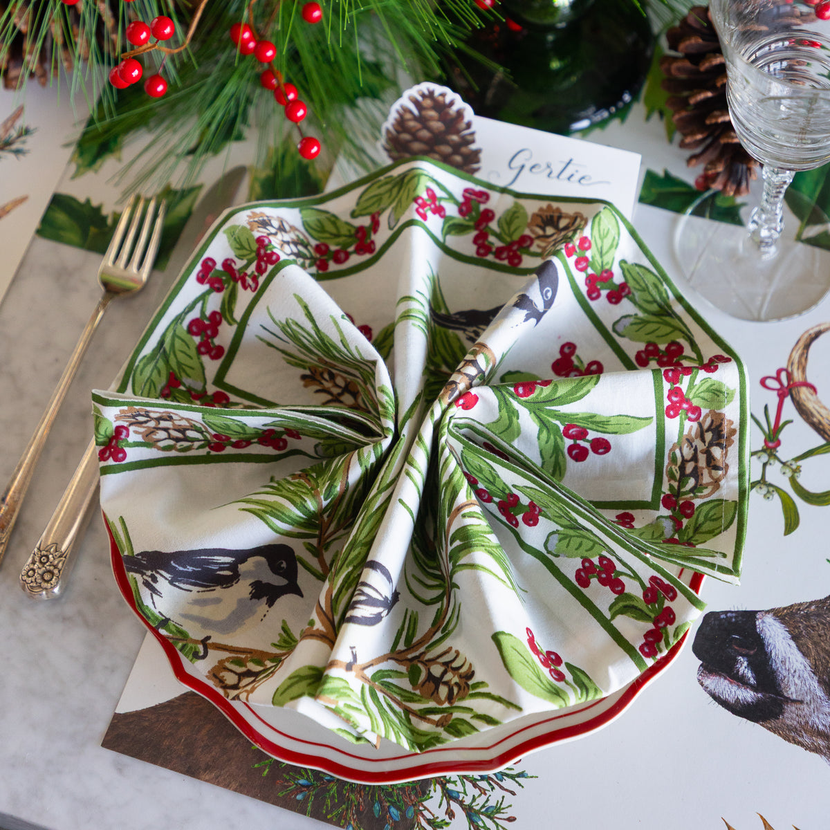 A folded white napkin with a woodland bird and berry print is arranged on a place setting table runner, surrounded by gold-toned flatware, pinecones, evergreen branches, and a glass.