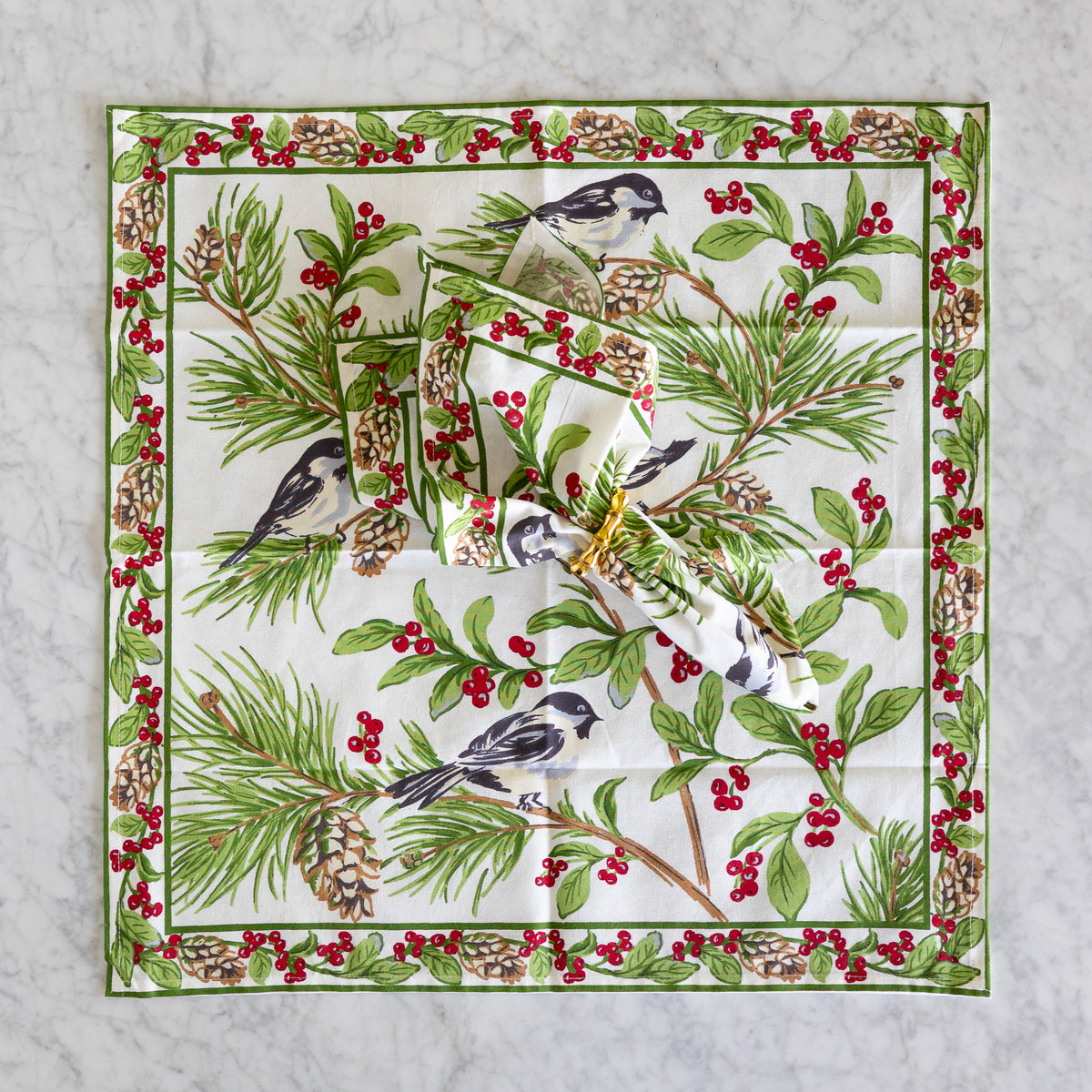 A square white fabric napkin with a detailed winter woodland print featuring birds, pine branches, pinecones, and red berries, folded and stacked on a light gray marble surface.
