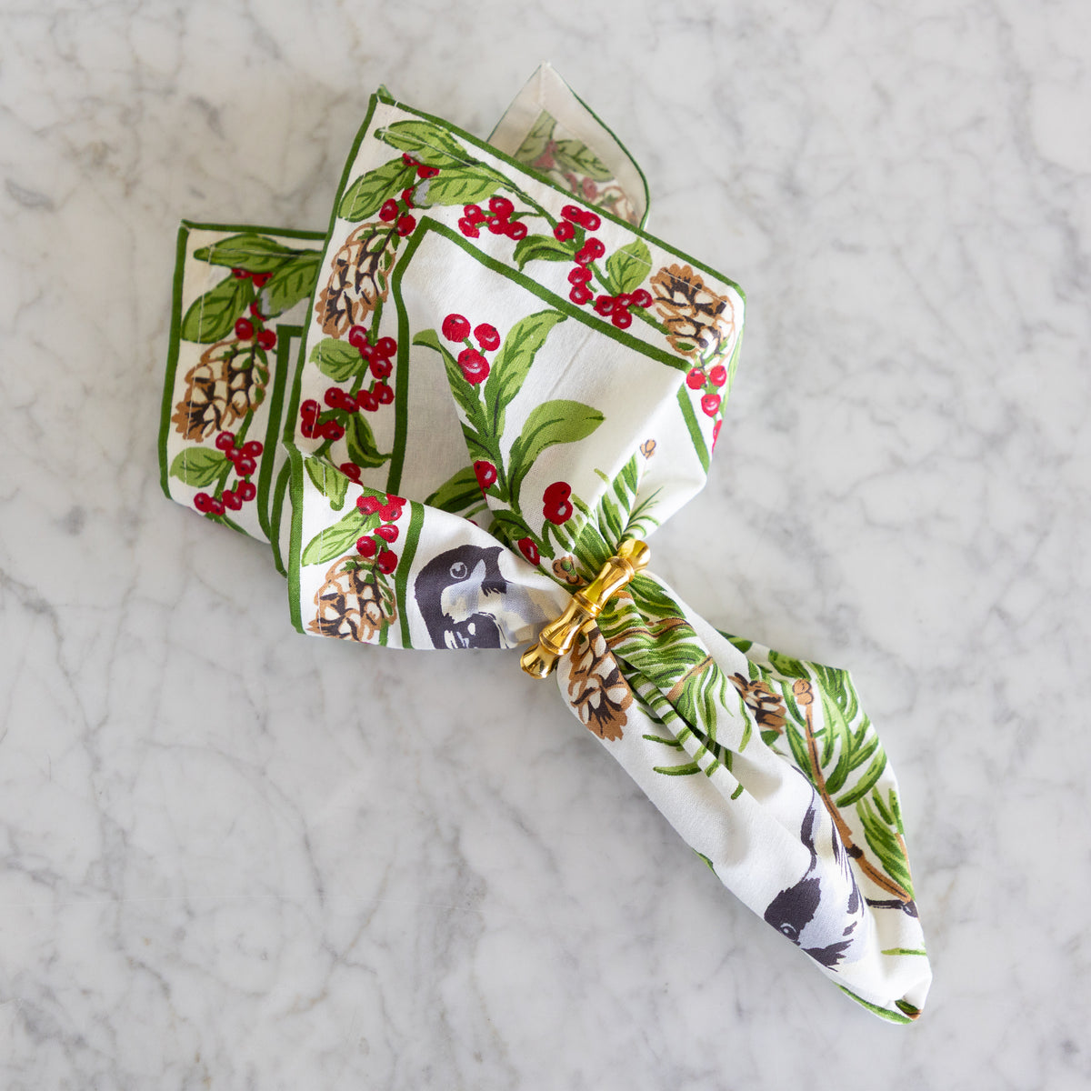 A square white fabric napkin with a detailed winter woodland print featuring birds, pine branches, pinecones, and red berries, folded and stacked on a light gray marble surface.