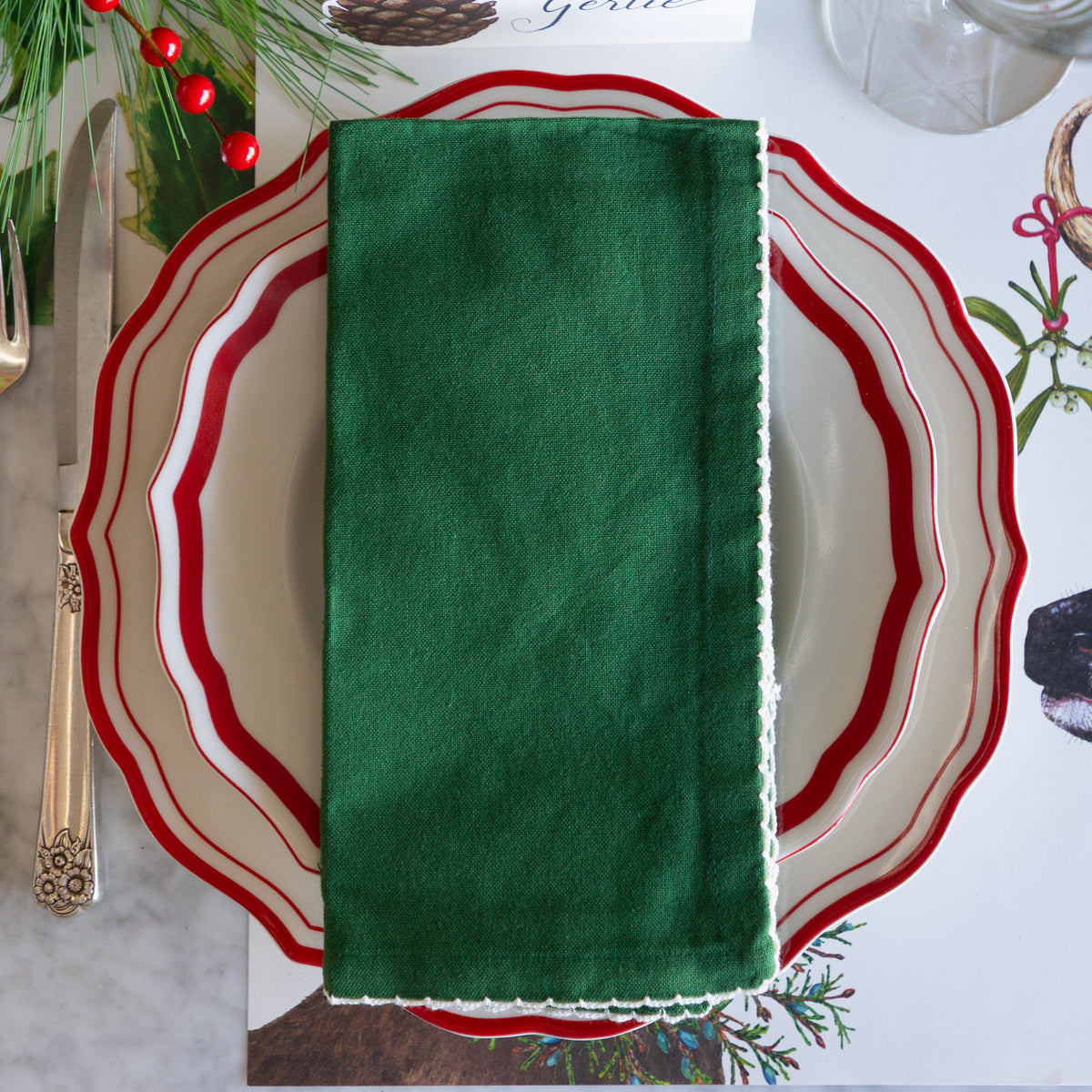 A dark green cotton napkin with white lace trim, neatly folded and placed on a white plate with red and white wavy borders, set on a holiday-themed table.
