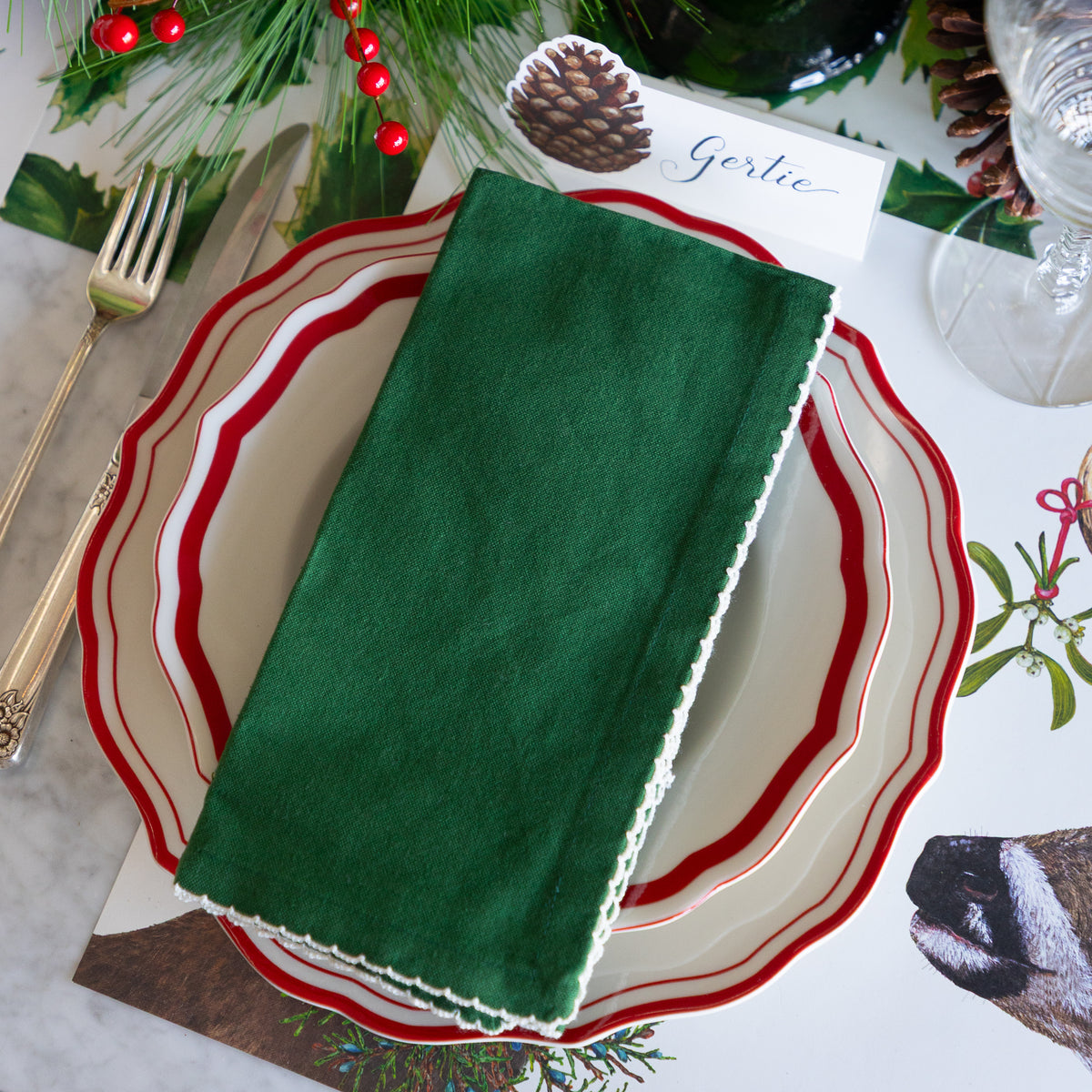 A dark green cotton napkin with white scalloped edges is folded and placed on a layered place setting of cream and red ceramic plates, styled with gold-toned cutlery, a pinecone name card, and holiday greenery.