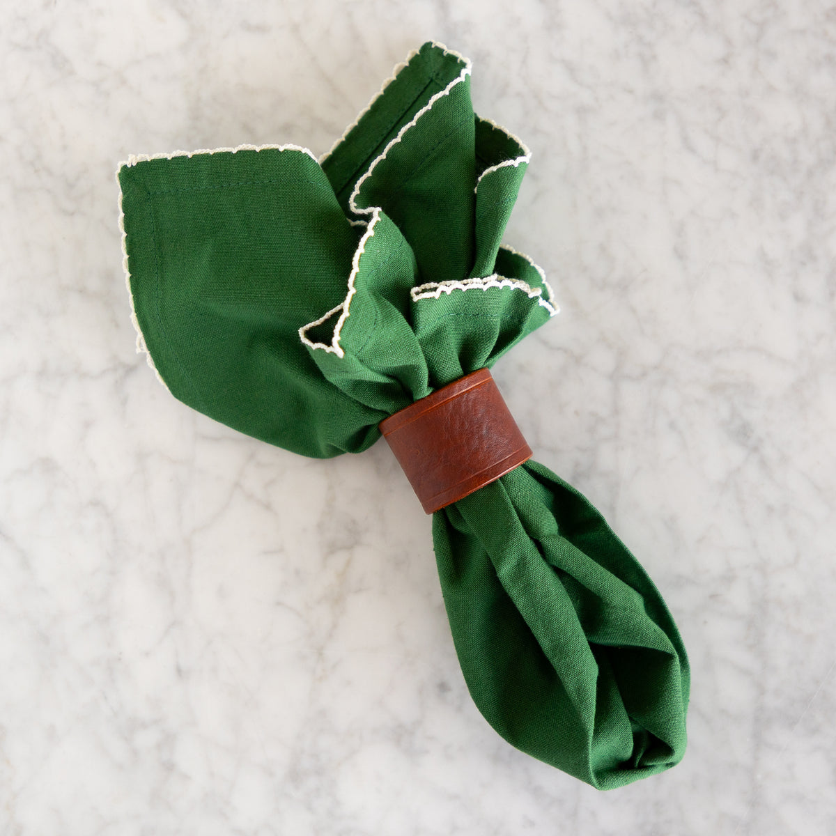 A dark green fabric napkin with white embroidered trim, gathered and secured with a brown leather napkin ring on a light gray marble surface.