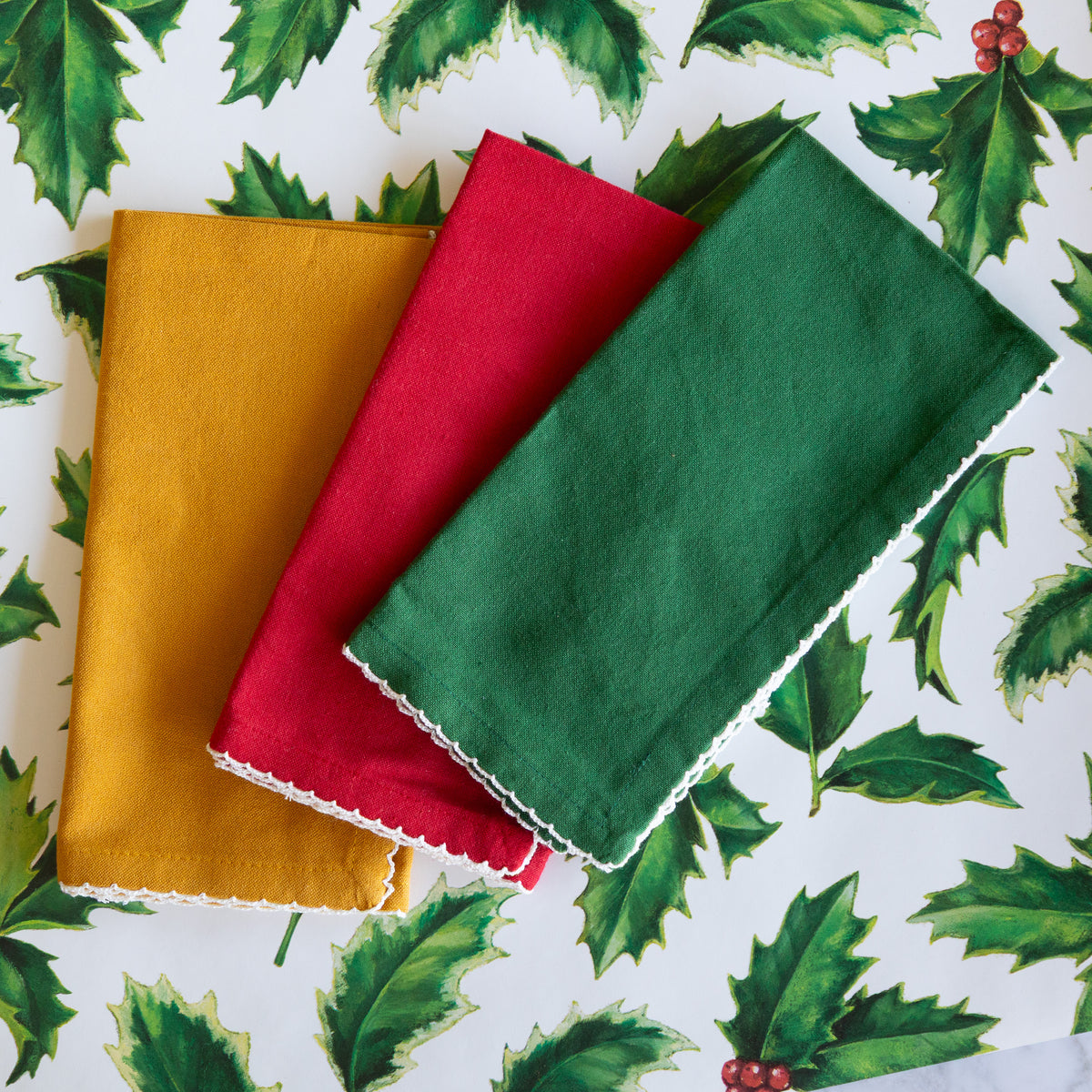 Three folded fabric napkins in mustard yellow, red, and green with white scalloped edges, arranged on a white background printed with green holly leaves and small red berries.