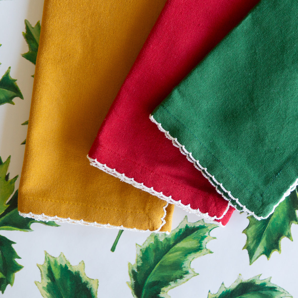 Three folded fabric napkins in mustard yellow, red, and green with white scalloped edges, arranged on a white background with green holly leaf print.