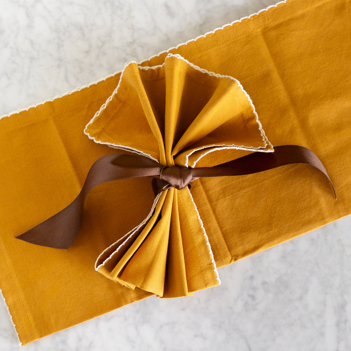 A folded mustard yellow fabric napkin with white scalloped edges, tied in a bow with a brown ribbon, placed on a white marble surface.