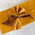 A folded mustard yellow fabric napkin with white scalloped edges, tied in a bow with a brown ribbon, placed on a white marble surface.